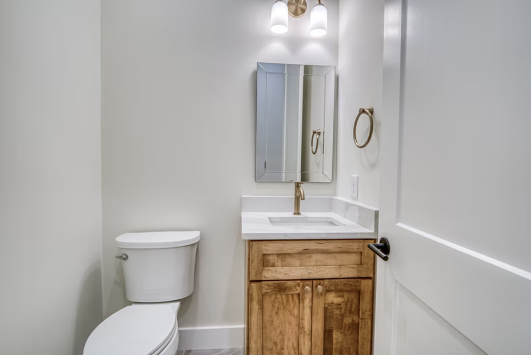 Bathroom with a toilet, wooden vanity with a marble countertop, a small mirror, a gold faucet, and a wall-mounted towel ring, with modern light fixture above.