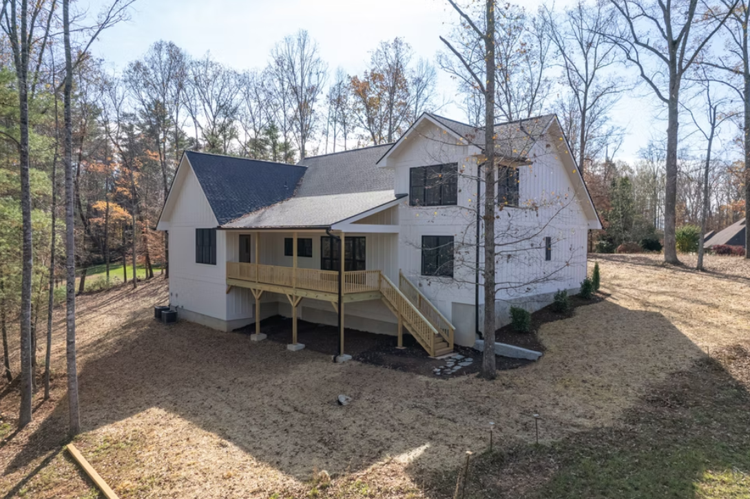 A two-story white house with a gray roof in a wooded area, featuring a wooden deck and staircase on the back over a yard with sparse grass and trees.