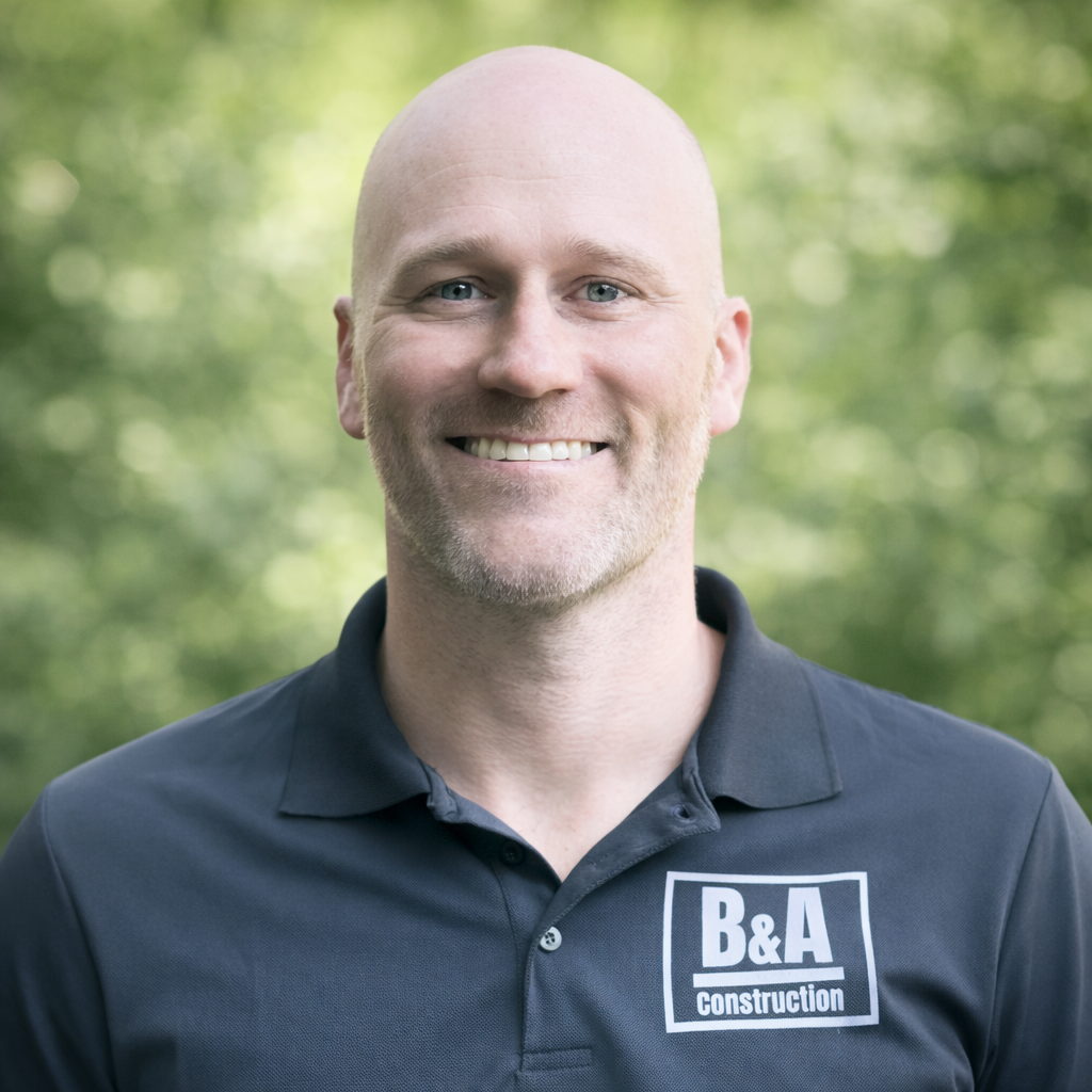 A smiling man with a bald head and blue eyes wearing a black polo shirt with a B&A Construction logo, standing outdoors with a blurred green foliage background.