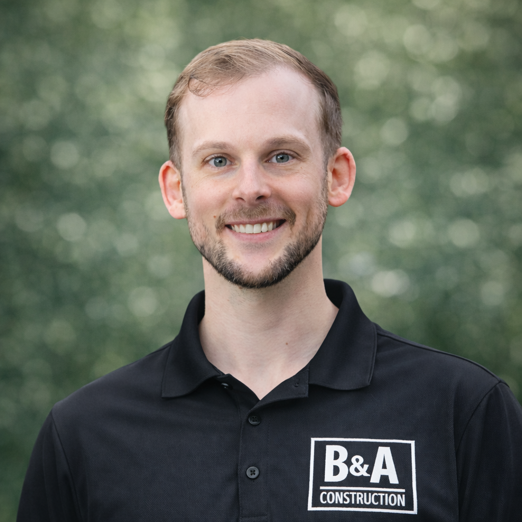 A young man with light skin, short light brown hair, and a beard, smiling outdoors, wearing a black polo shirt with a 'B&A Construction' logo.