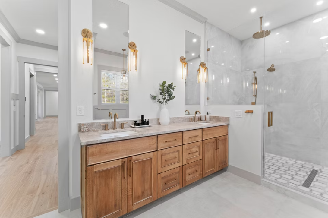 Modern bathroom with double vanity, wooden cabinets, marble countertop, large mirror, pendant lights, white walls, and a glass walk-in shower with marble tiles and a rain showerhead.