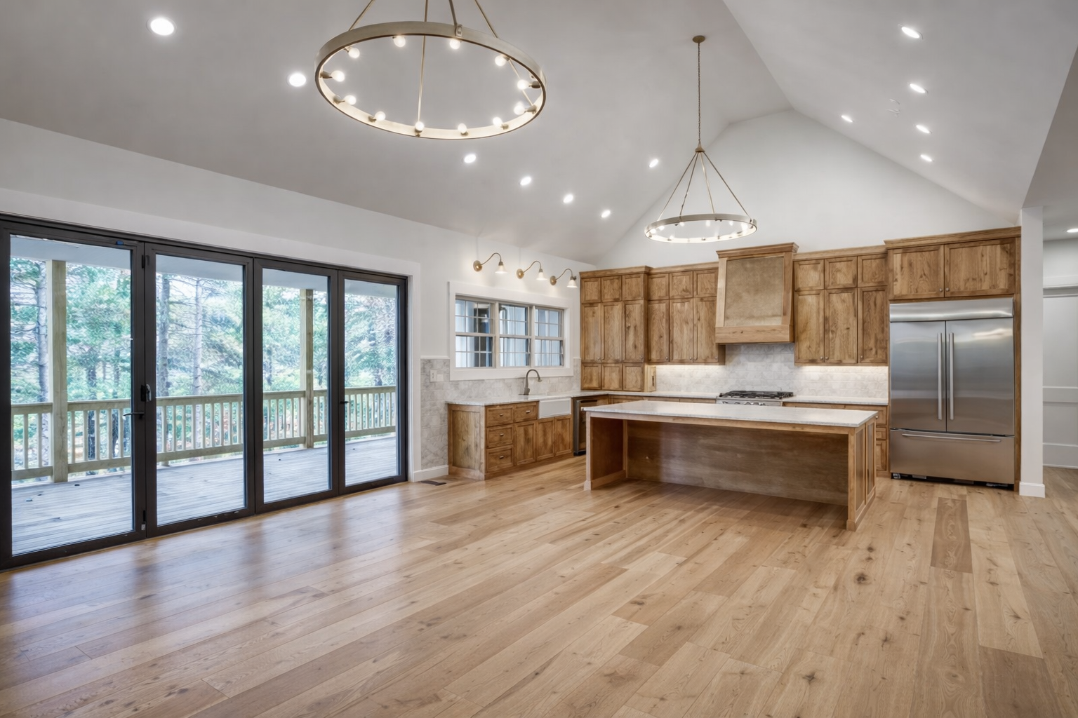Open-concept kitchen with wooden cabinets, a kitchen island, and large sliding glass doors leading to a deck, with a view of a wooded area outside. Recessed ceiling lights and two circular chandeliers illuminate the room.
