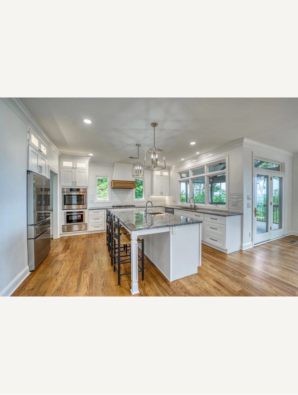 Bright kitchen with white cabinets, granite countertops, hardwood floors, built-in oven, microwave, stovetop, large island with seating, multiple windows, and a glass door leading to an outdoor deck.