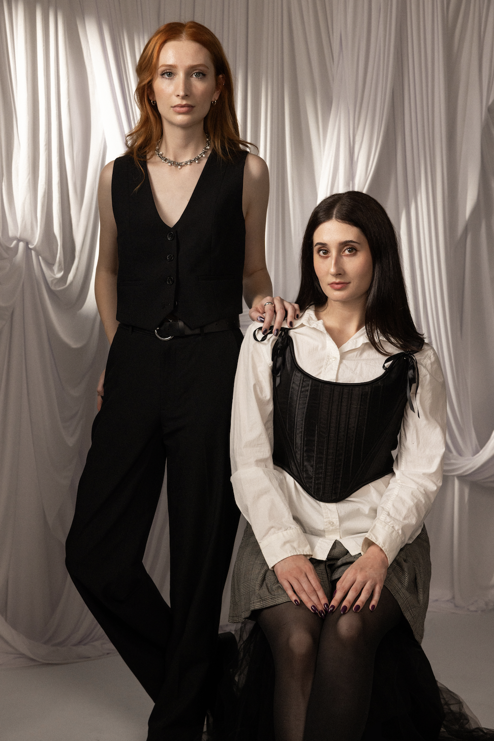 A tall person with long red hair standing next to a woman with long brown hair sitting in a chair against a white backdrop.