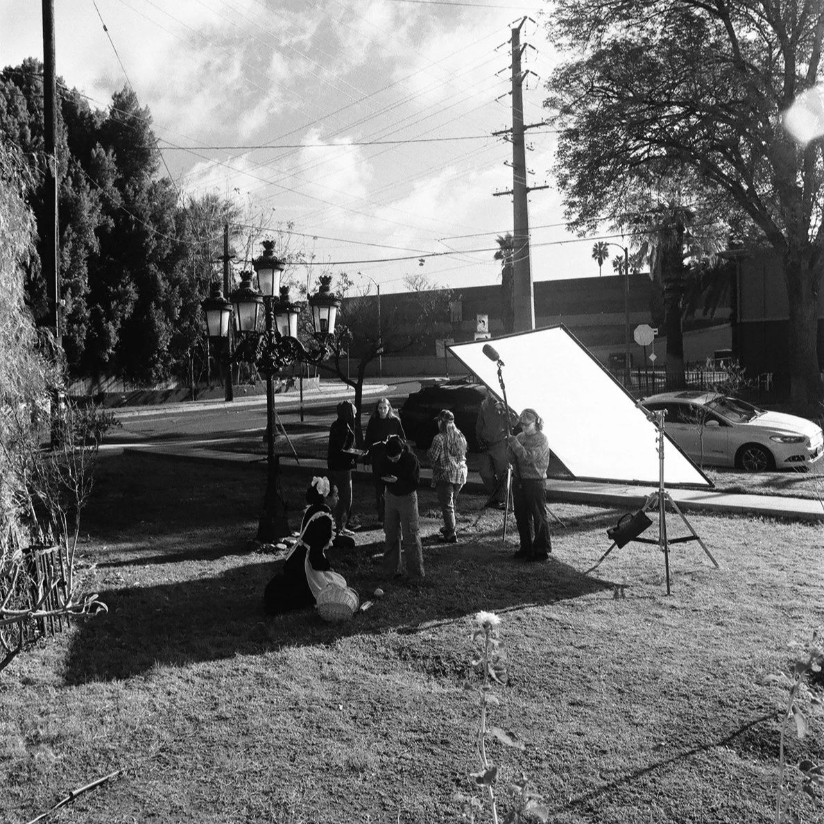 Black-and-white photo of a film crew setting up a scene outdoors with a large reflector and lighting equipment. Several people are gathered around, some standing and others kneeling, with one person in historical costume. Trees, cars, a streetlamp, and utility poles are visible in the background.