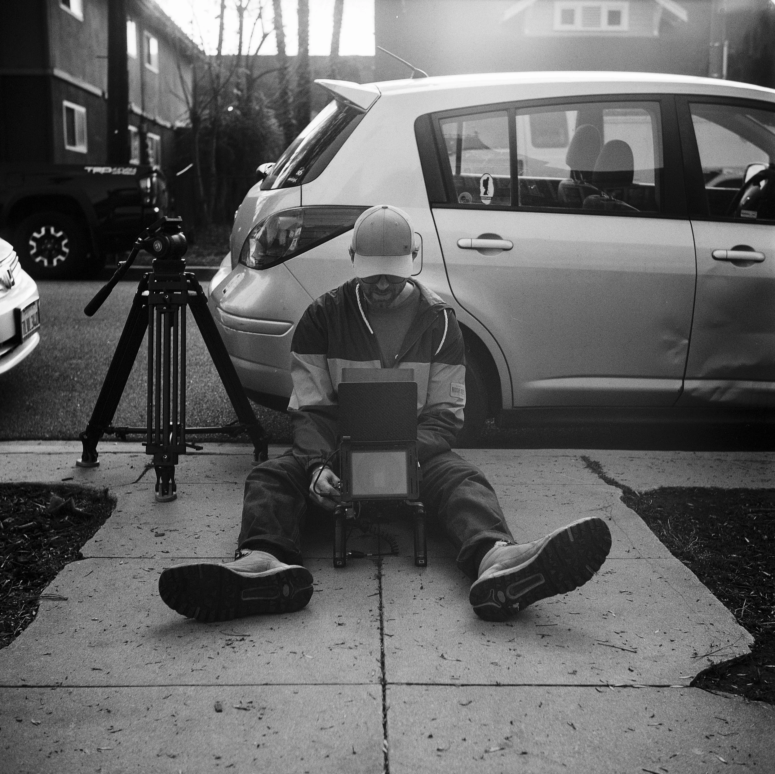 A person wearing a cap and outdoor clothing sitting on the sidewalk operating a camera or electronic equipment, with a tripod nearby, in front of parked cars on a residential street.