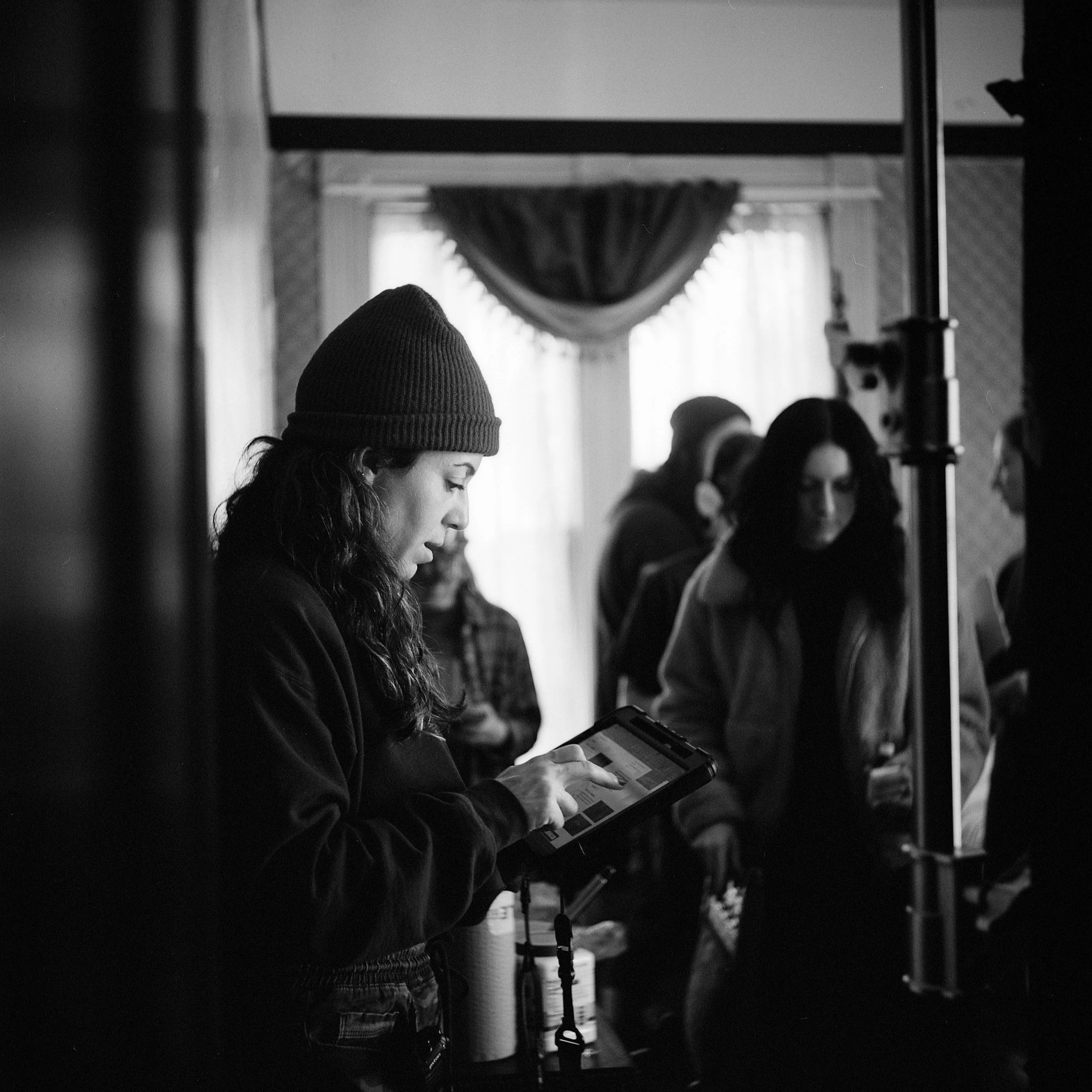 A woman wearing a beanie is looking at a tablet in a room with multiple people and a curtain in the background.