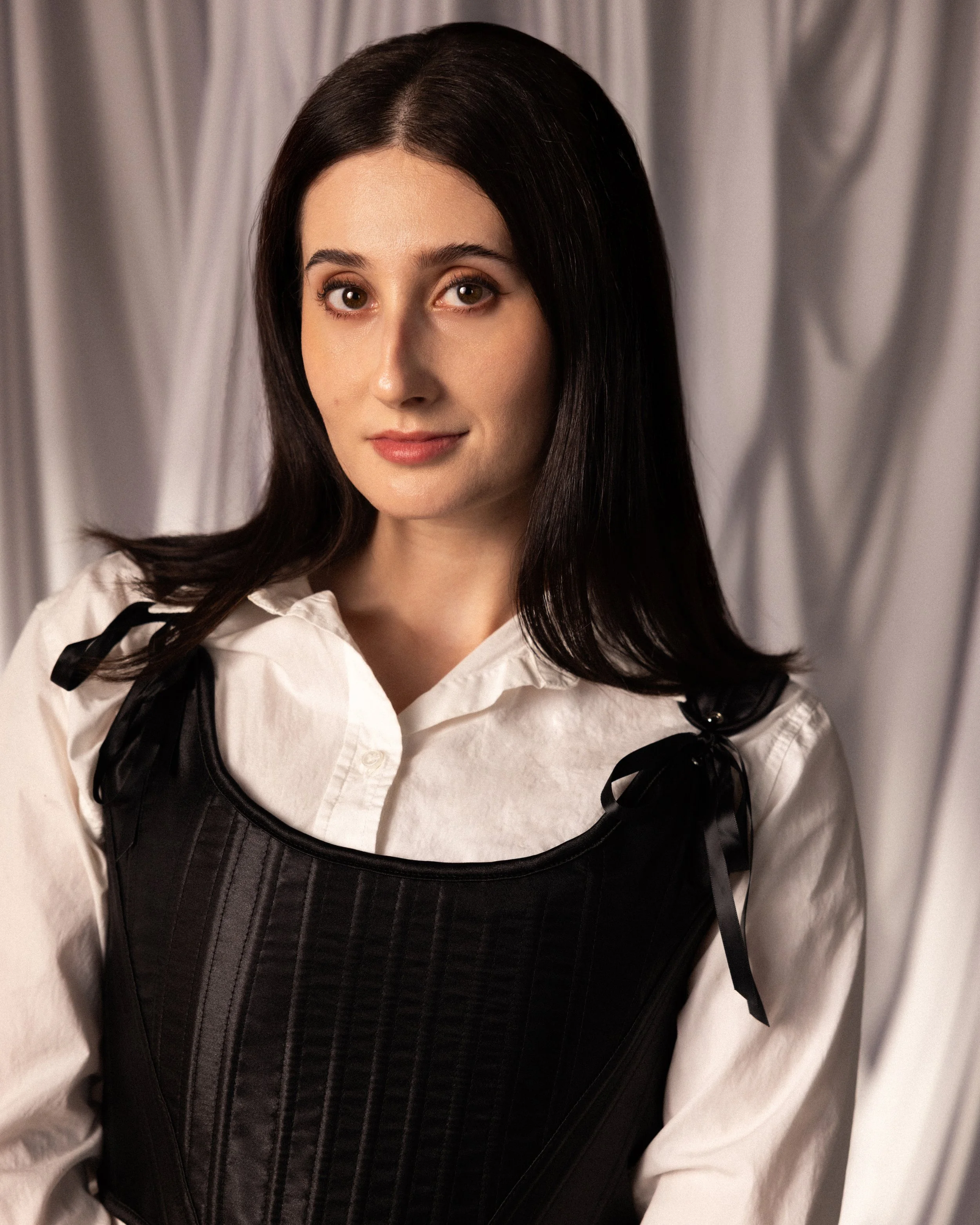 A woman with dark brown hair wearing a white shirt and a black corset.