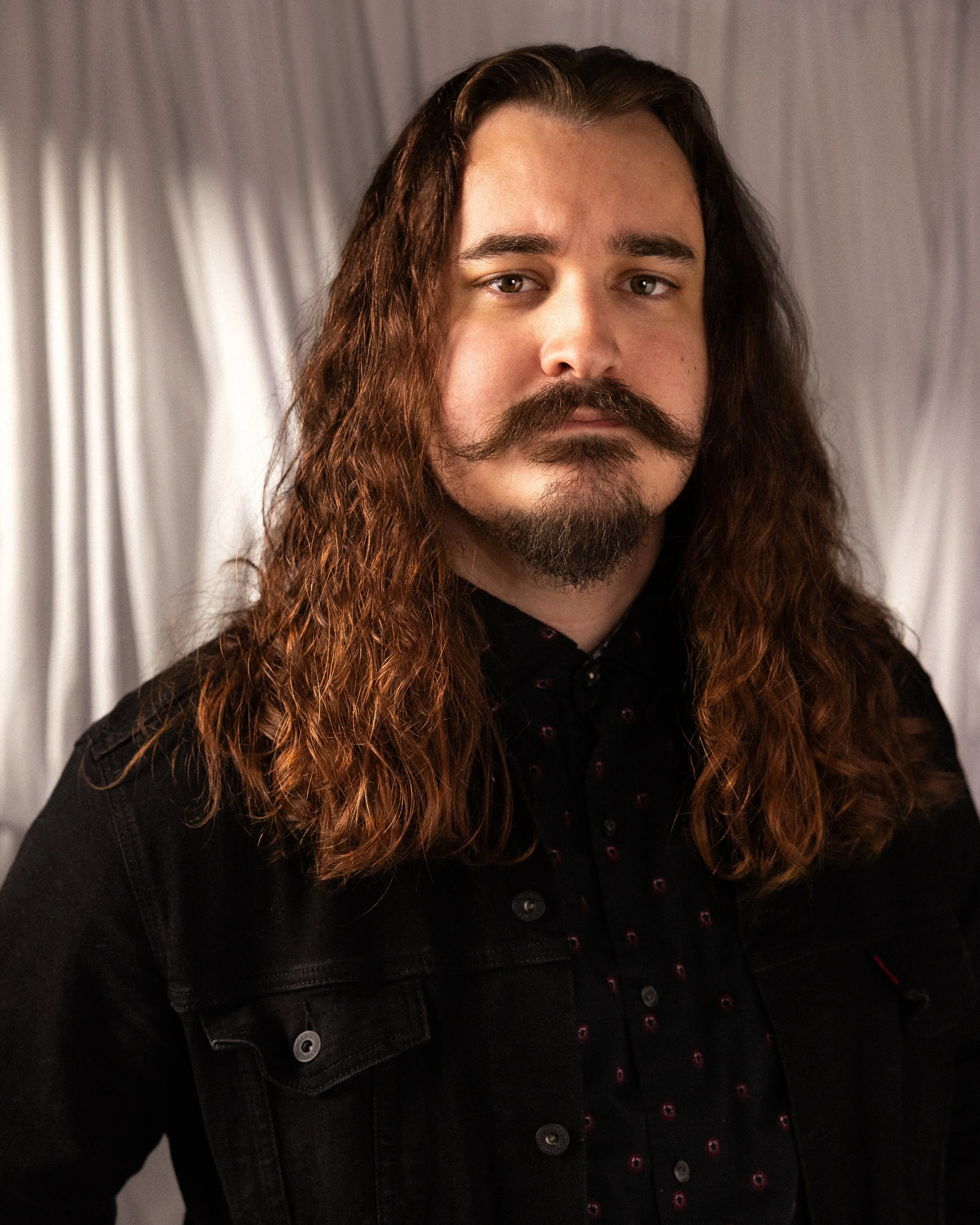 A man with long brown hair and a mustache wearing a dark shirt.