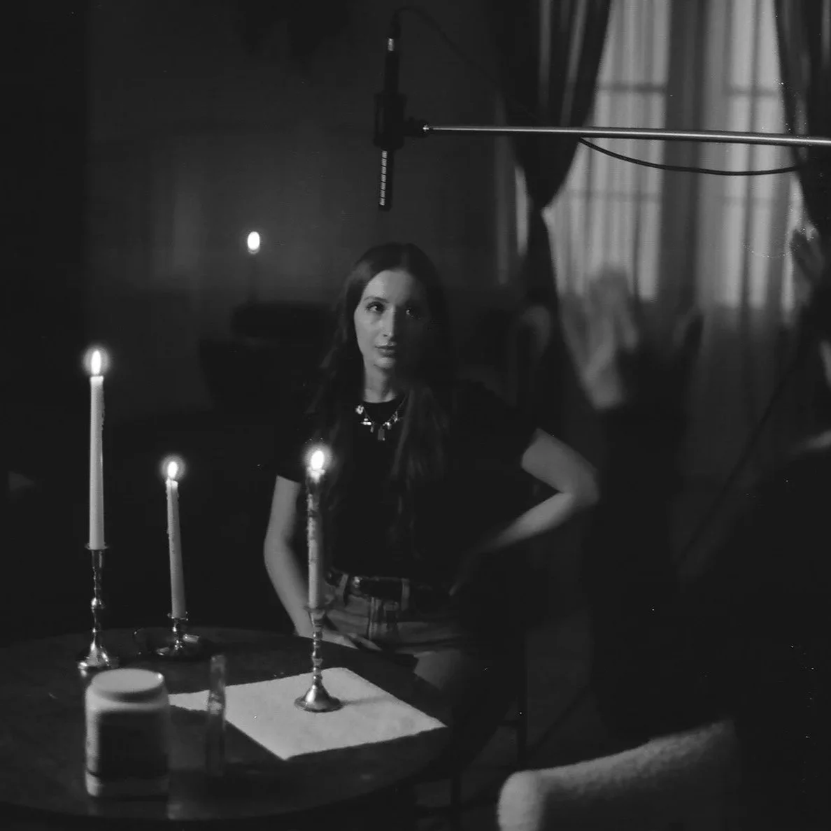 A woman with long dark hair sitting at a table with three lit candles, in a dimly lit room with curtains and a window in the background, being filmed with a microphone overhead.