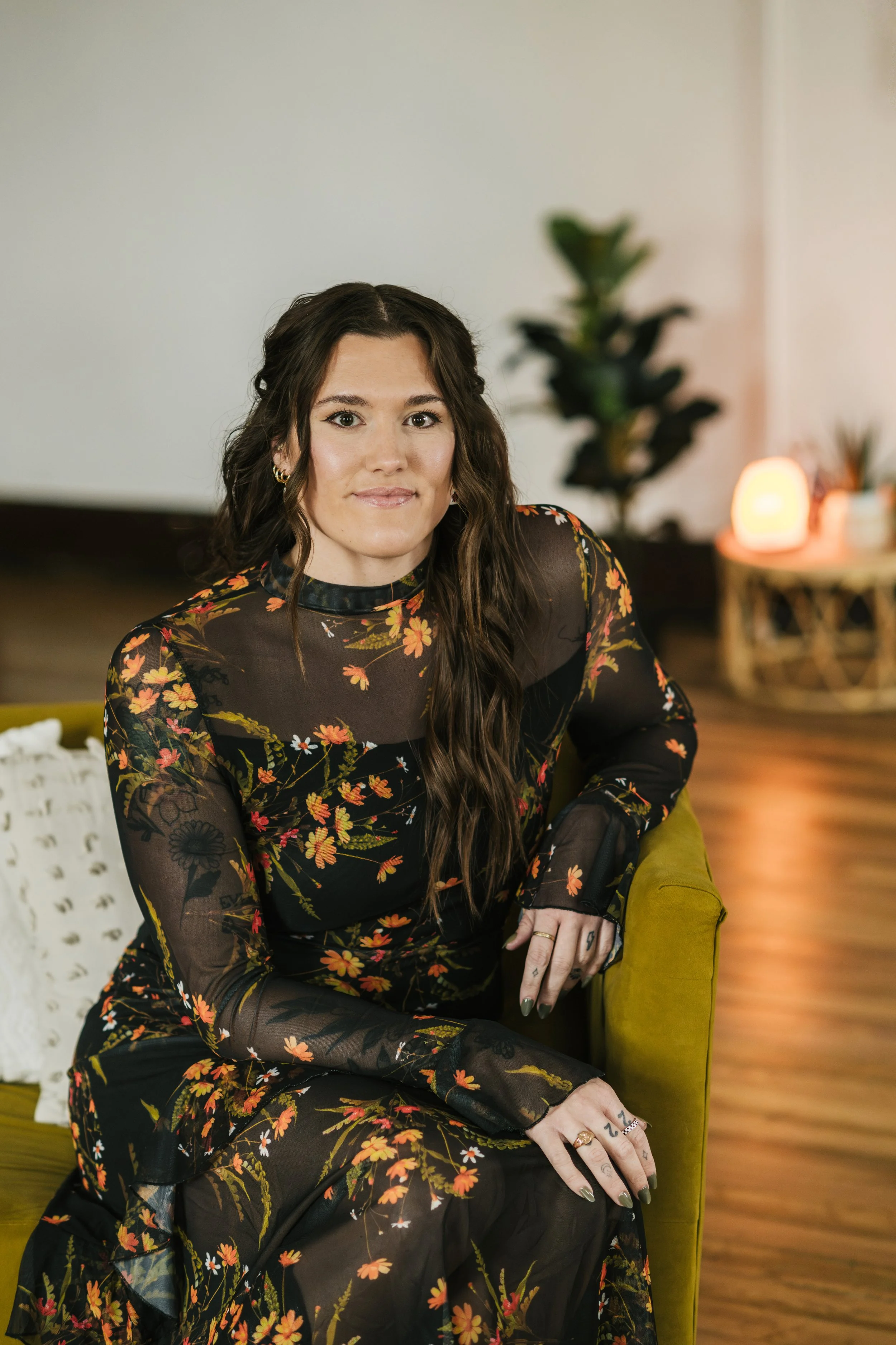 A woman with long wavy brown hair wearing a black sheer floral dress sitting on a green chair with a white pillow. In the background, there is a potted plant and a side table with a glowing lamp.