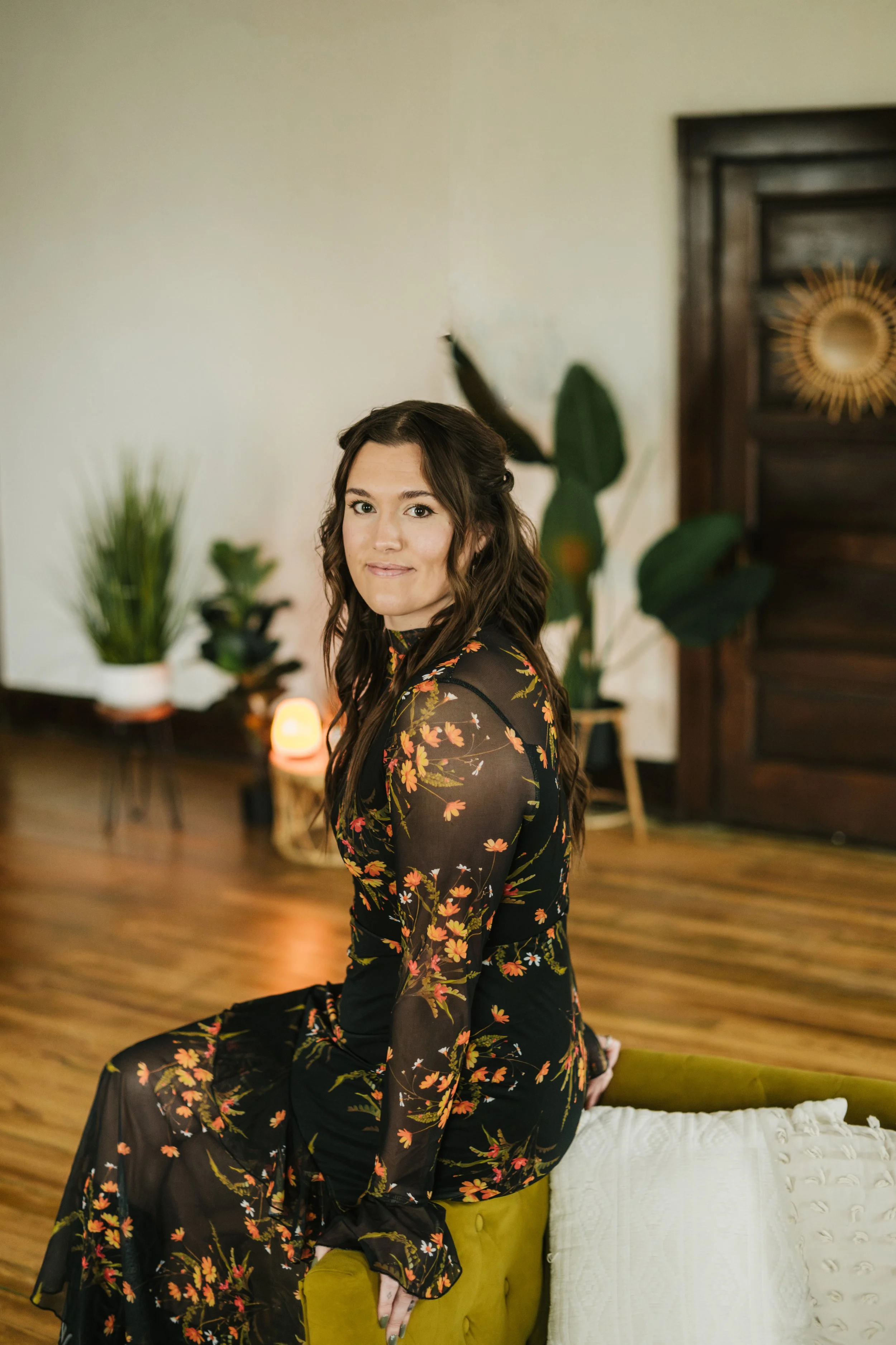 A woman with long dark hair wearing a black, floral-printed dress, sitting on the edge of a green couch in a living room with wooden floors and potted plants in the background.