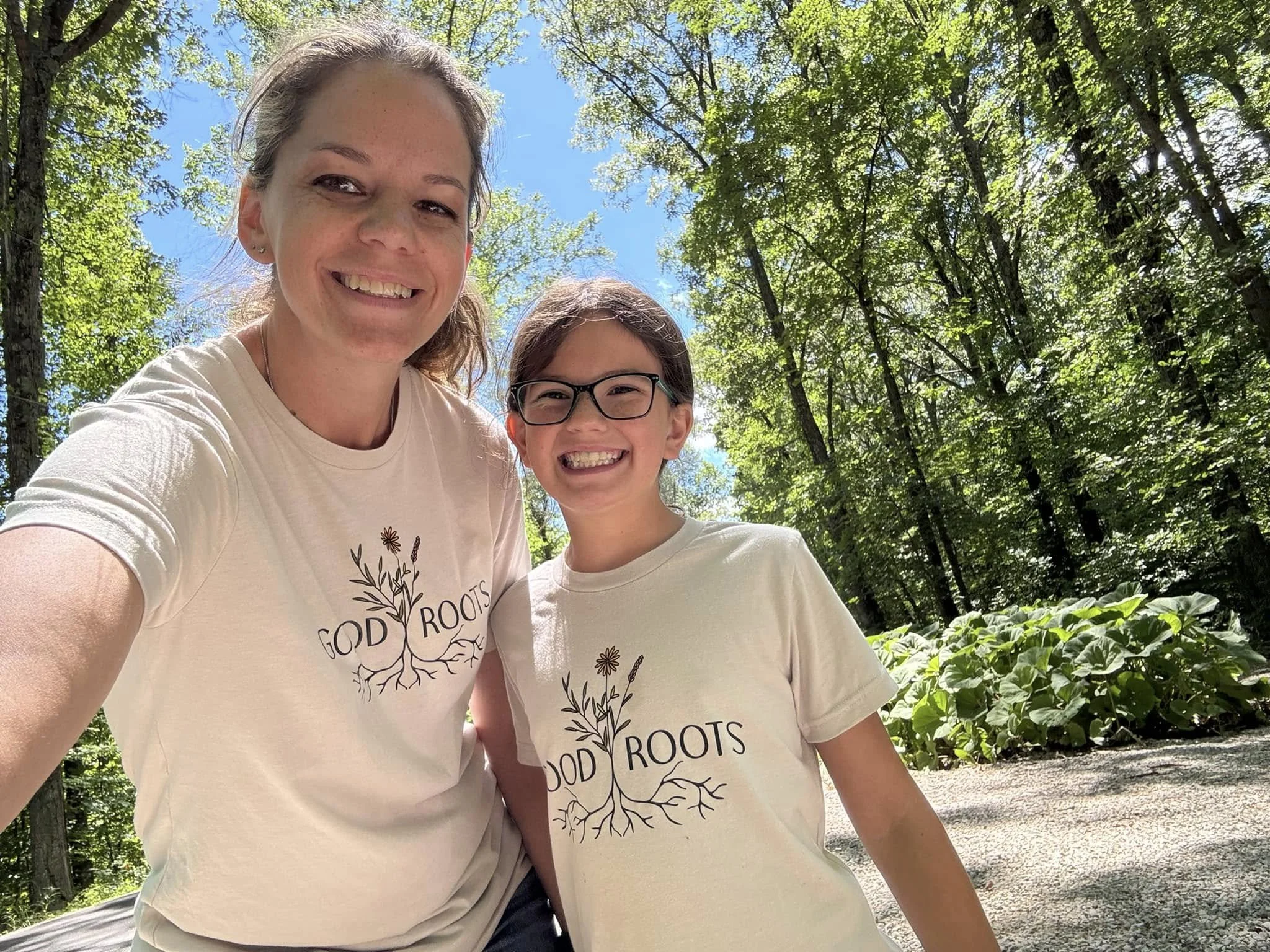 A woman and child smiling outdoors in a wooded area, both wearing matching T-shirts with 'GOD ROOTS' and a tree design.