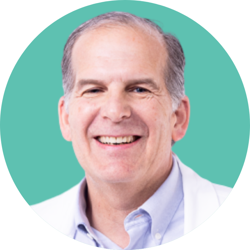 Portrait of a middle-aged man with short gray hair, smiling, wearing a light blue collared shirt and a white coat, against a teal background.
