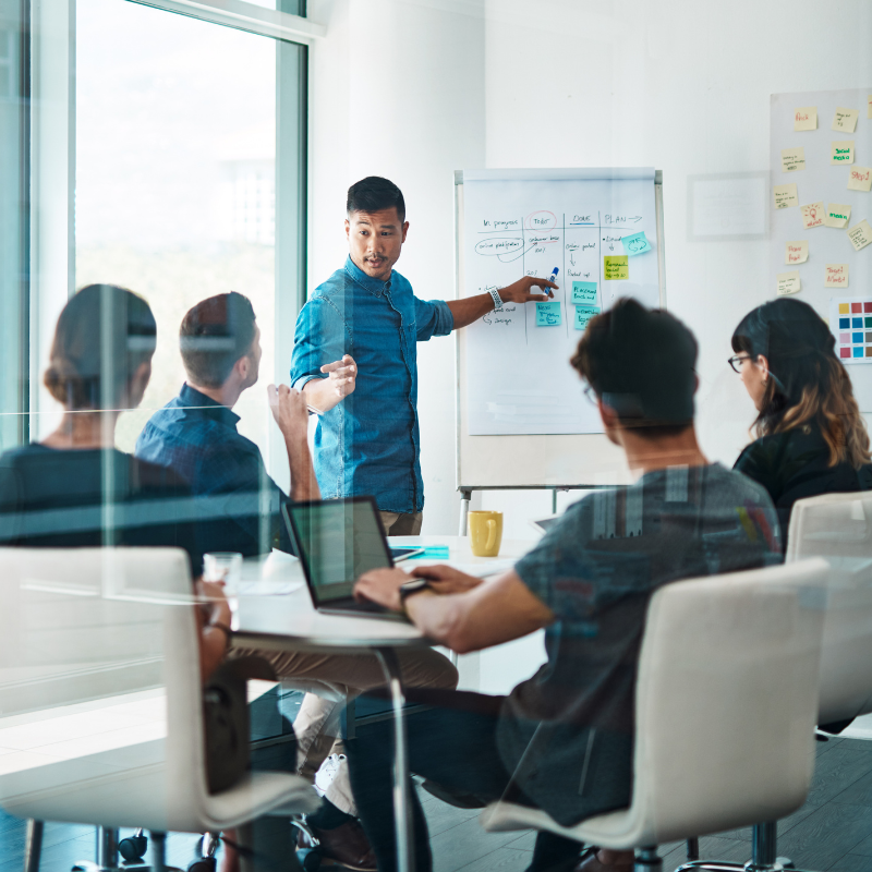 Man presenting to a small business meeting in a conference room with large window, using a whiteboard with notes and sticky notes.