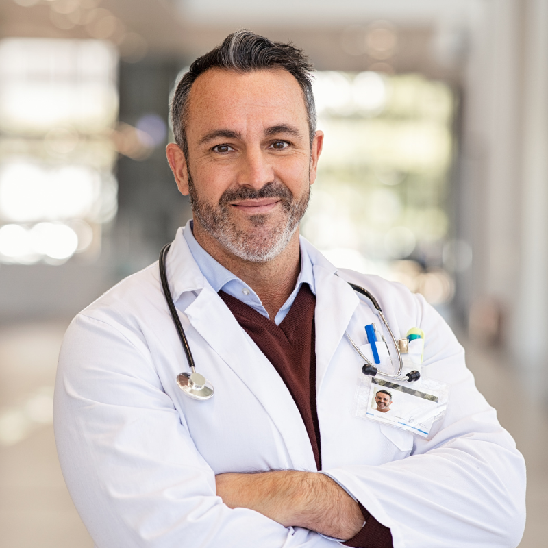A confident male doctor poses with arms crossed in a modern, well-lit hospital or clinic hallway, wearing a white lab coat with stethoscope and ID badge.