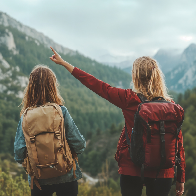 Two girls with backpacks hiking in a mountainous area, one pointing towards the mountains in the distance.
