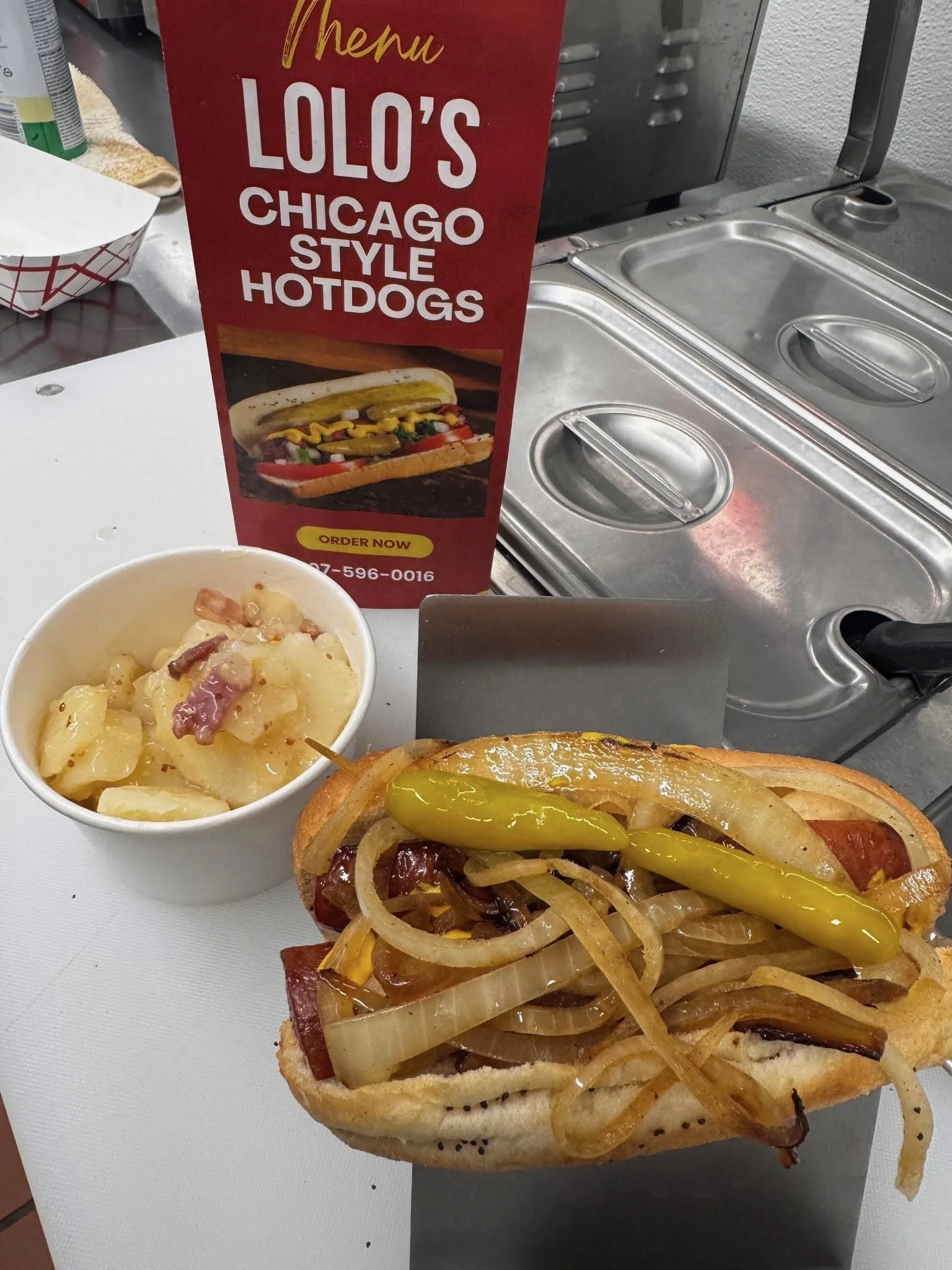A Chicago-style hot dog topped with onions, pickles, and mustard, accompanied by a side dish of potato chips, and a sign advertising Chicago style hot dogs from Lolo's in the background.
