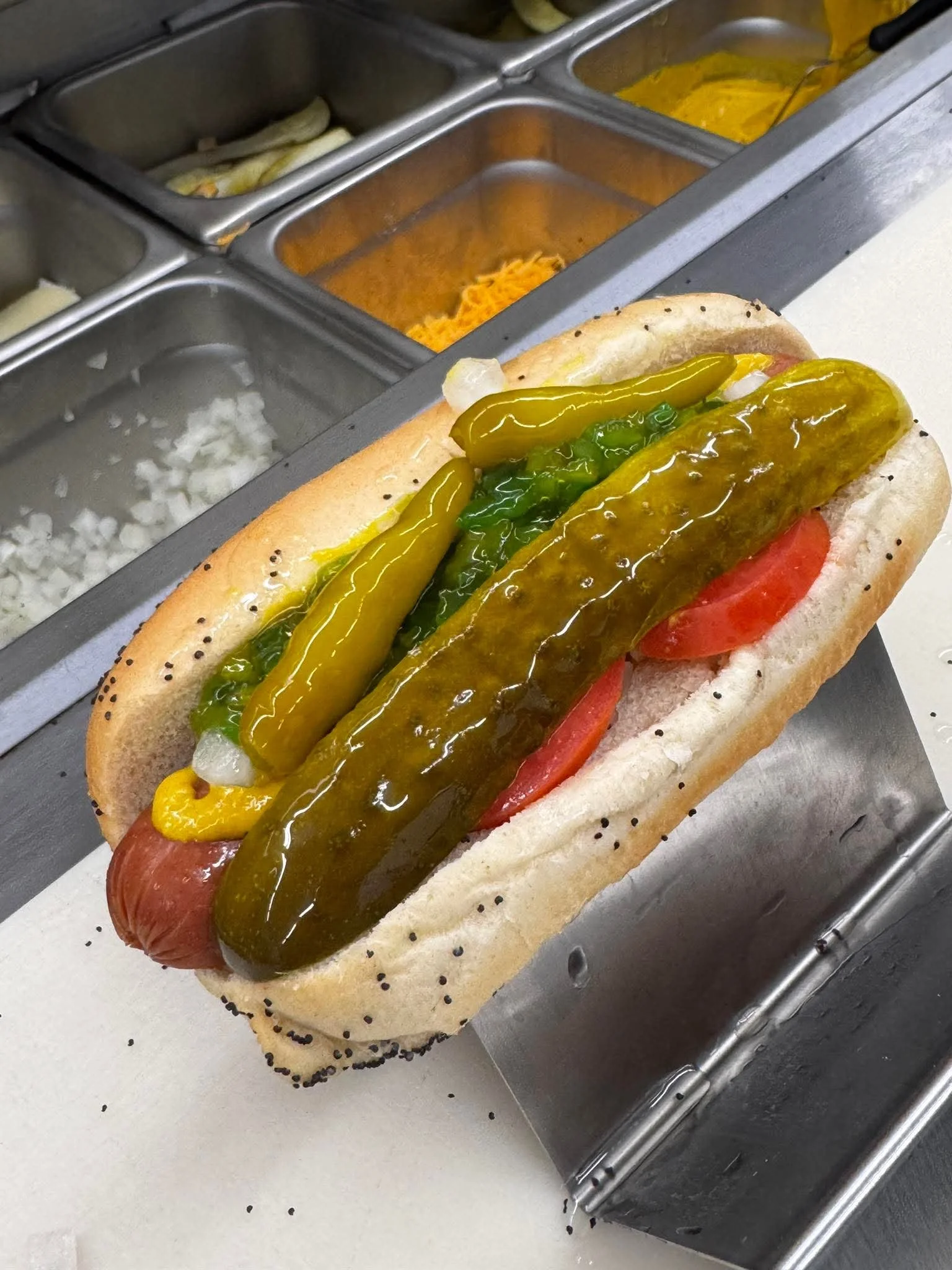 Hot dog with mustard, relish, pickles, onions, tomato slices, and poppy seed bun.