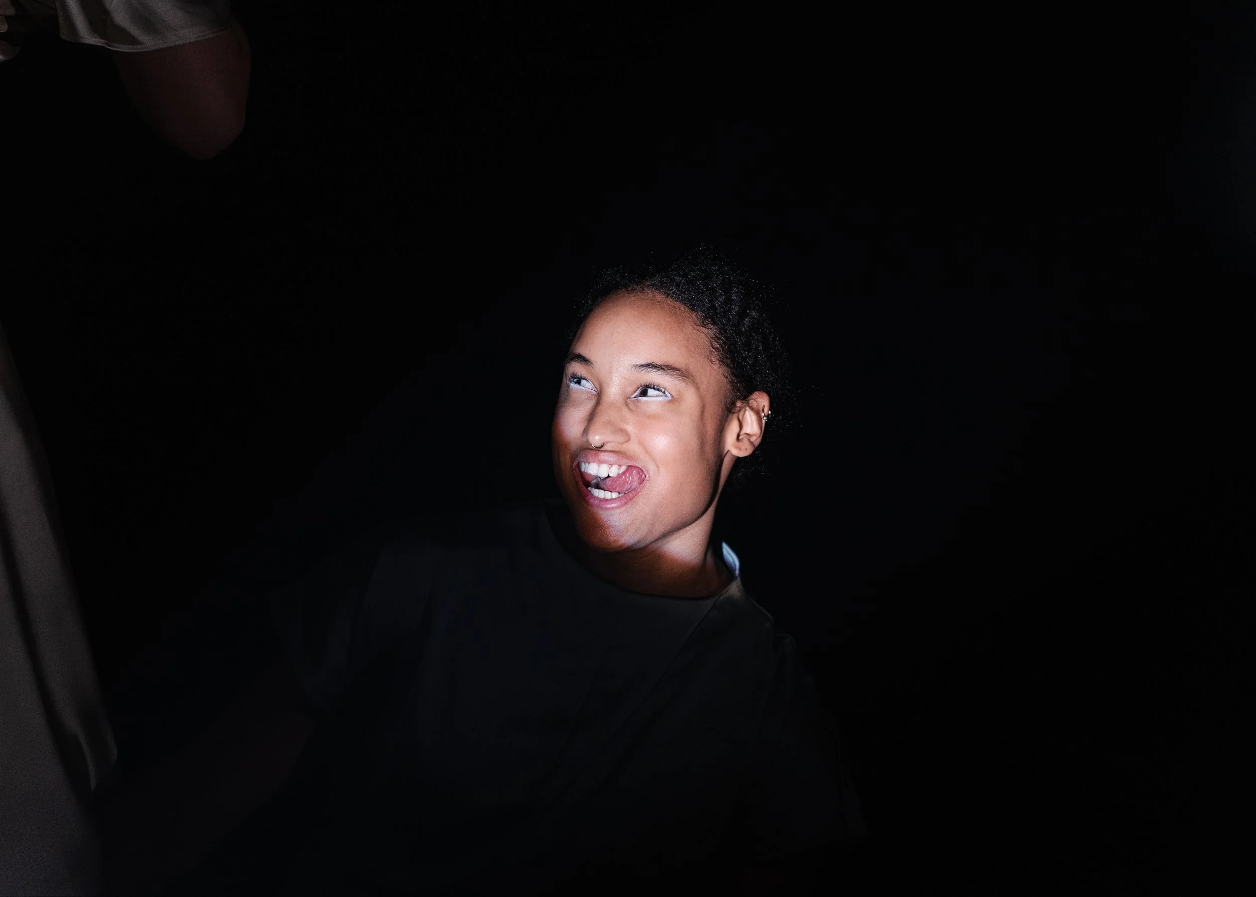 A young person with curly hair and a septum piercing making a funny face, sticking out their tongue and rolling their eyes, against a dark background.
