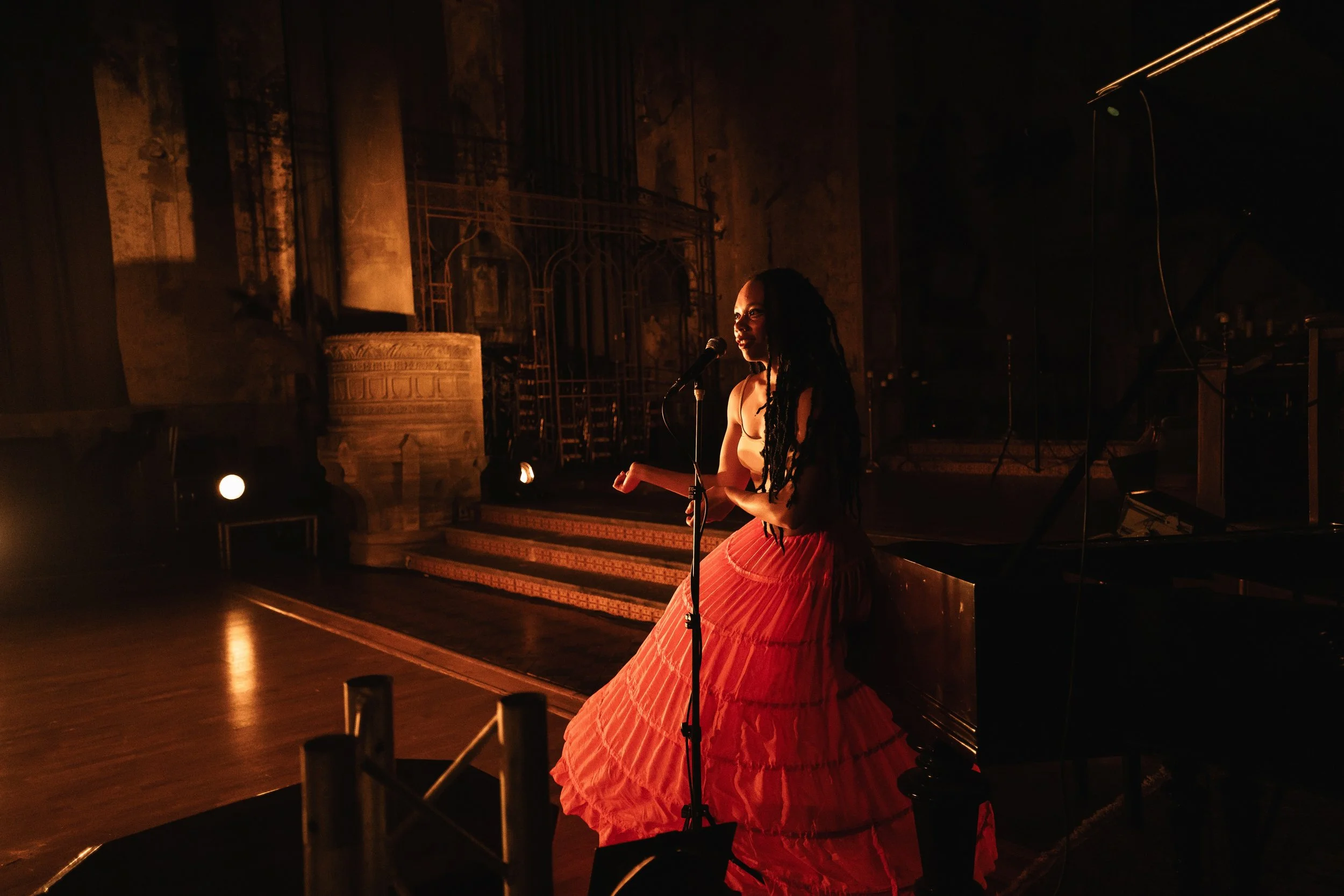 A woman with dreadlocks is on stage, singing into a microphone, wearing a long, crinoline orange skirt and a beige top, illuminated by warm stage lighting, with a grand piano nearby and an ornate, historic background.