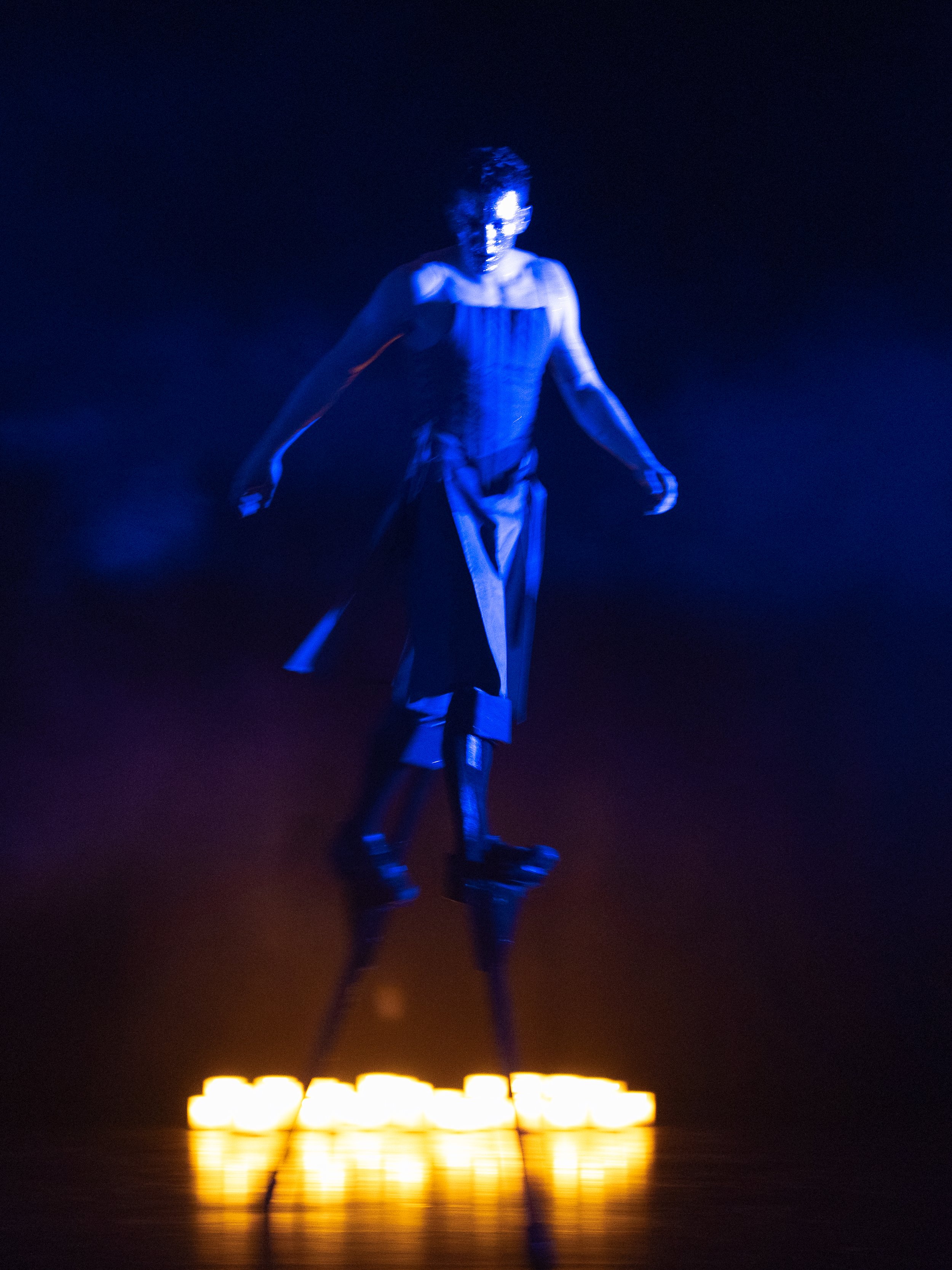 A person dressed in black, walking on stilts, illuminated by blue and yellow lights in a dark setting.