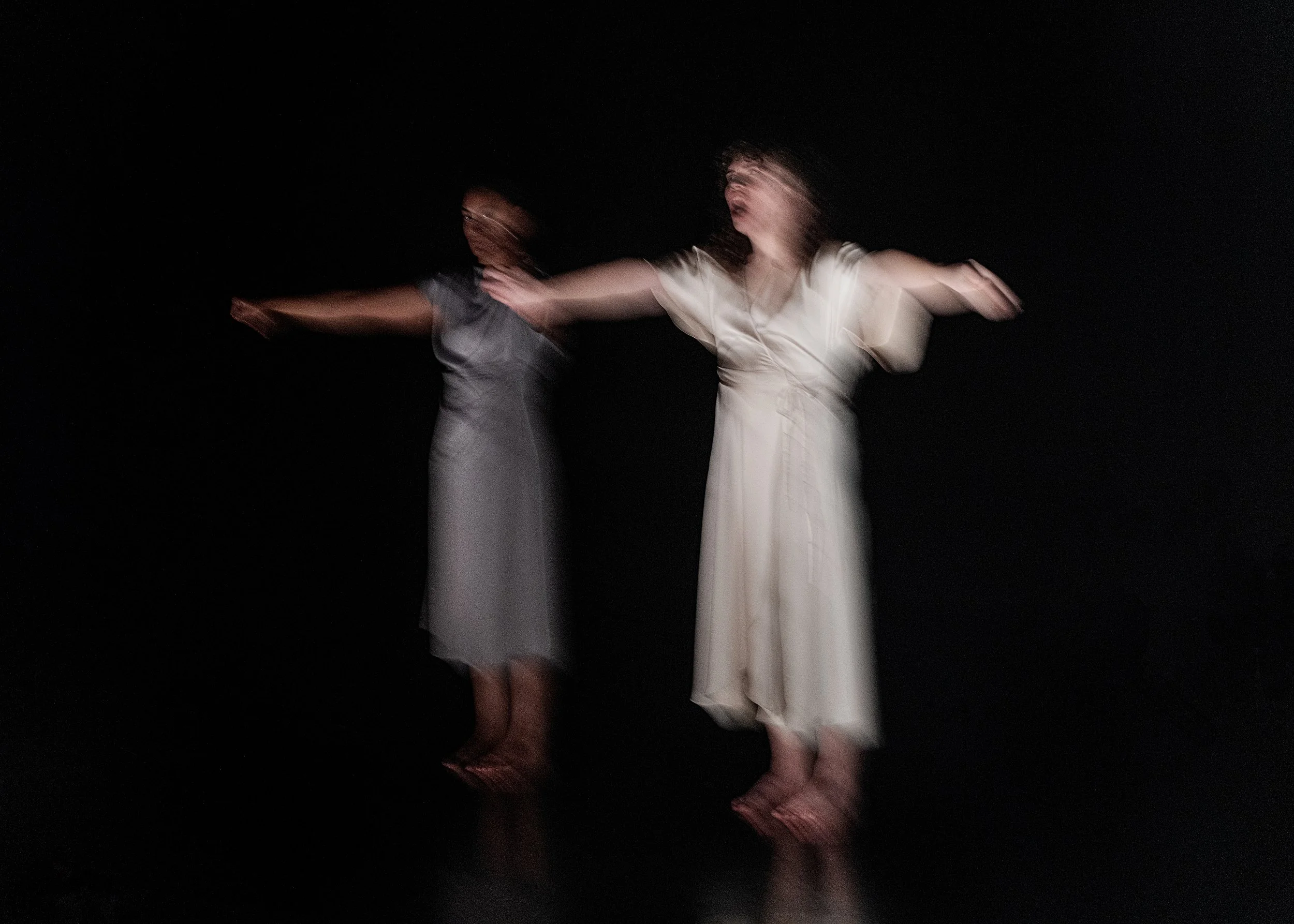 Two women in long dresses are dancing with outstretched arms in a dark space, captured with motion blur.