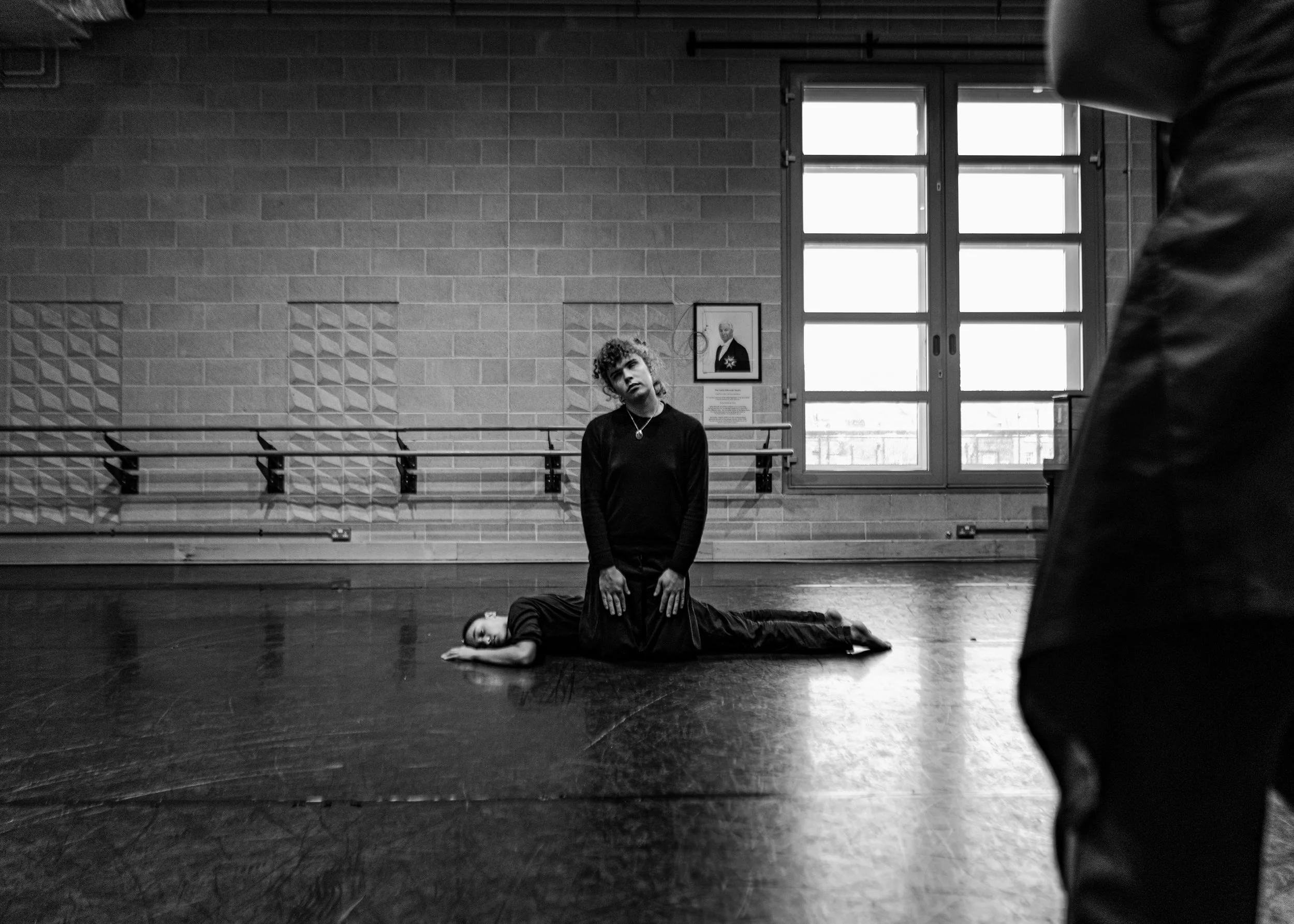 A dancer lying on their side on the floor with a woman kneeling on their back in a dance studio.