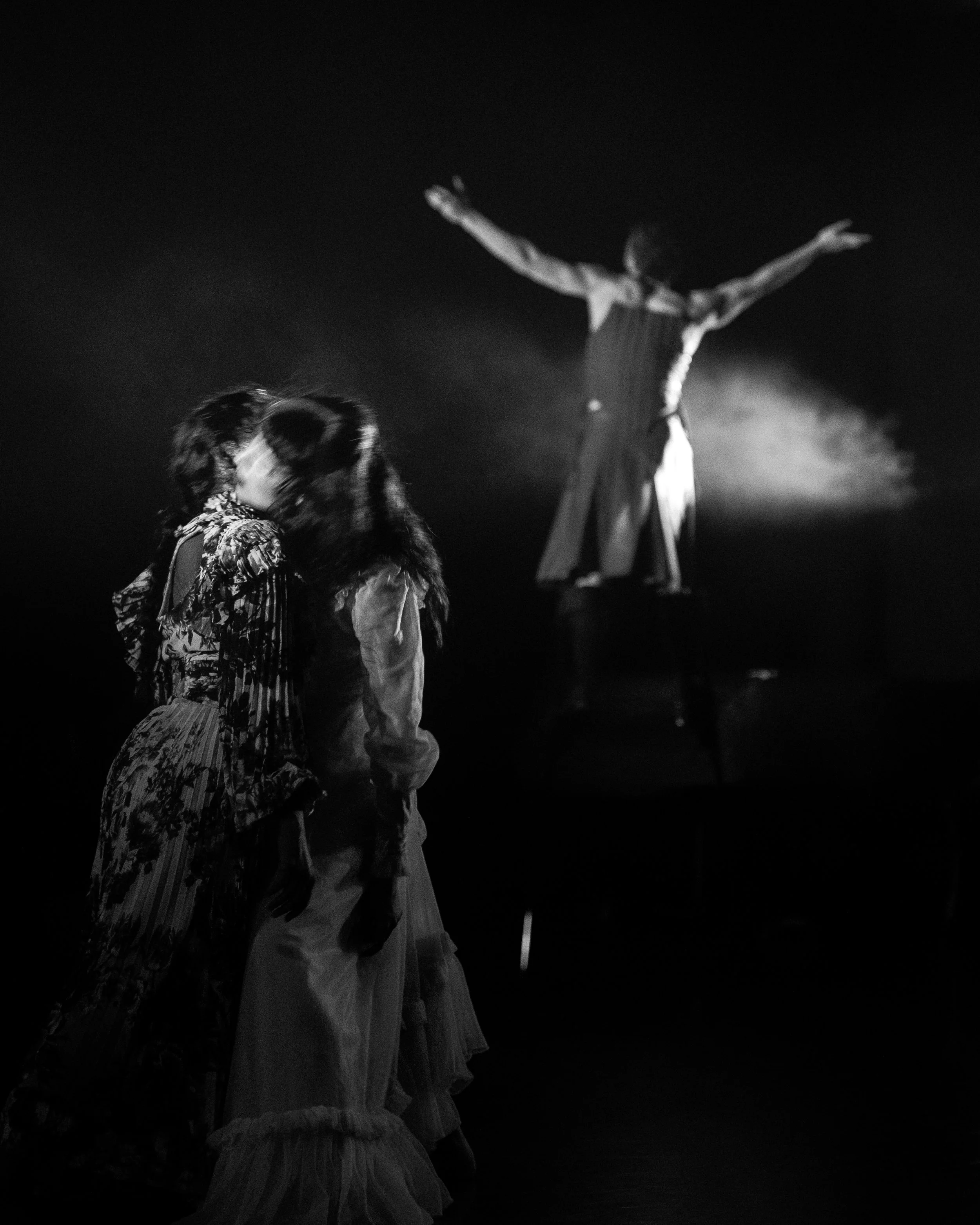 Two women in vintage clothing wrapping their necks around one another in the foreground, a person in period costume standing with arms outstretched in the background, all in black and white.