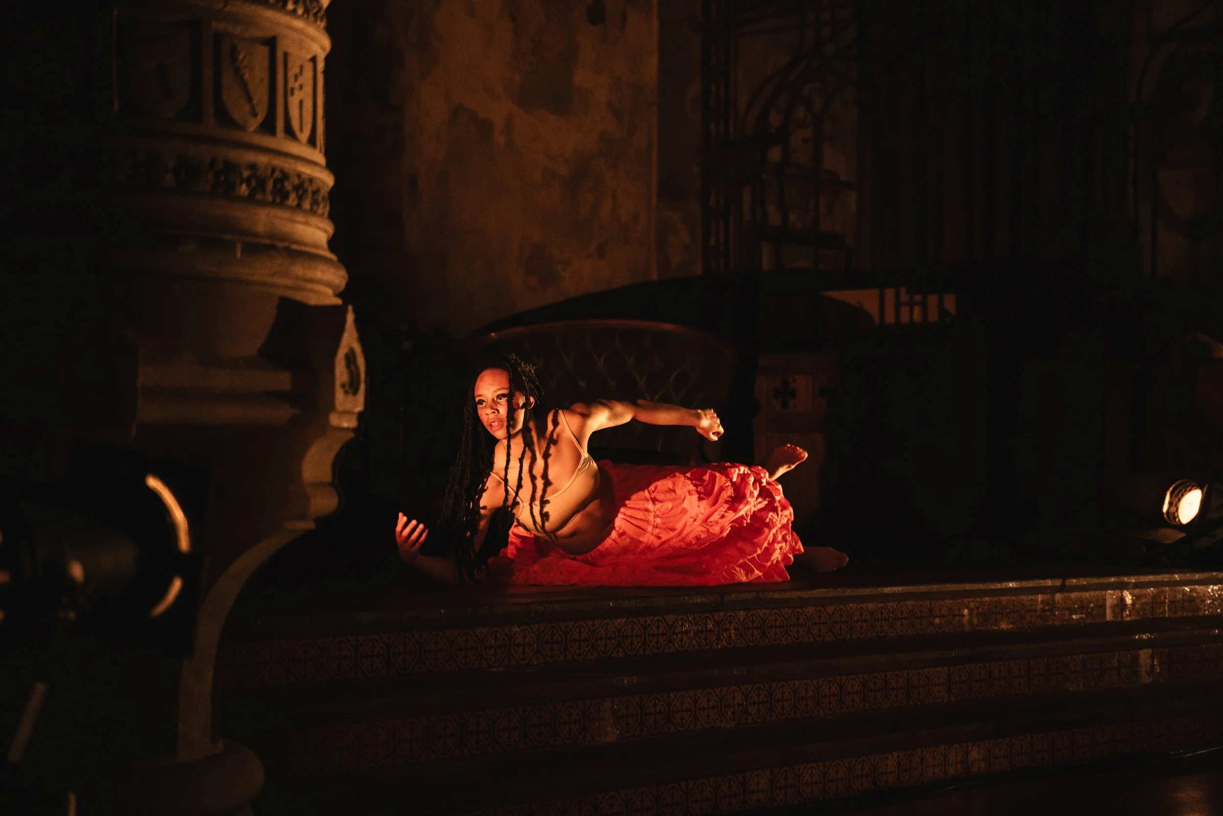A woman with light brown skin wearing a skin tone top lying on a stage, illuminated by a spotlight, with ornate architectural details in the background.