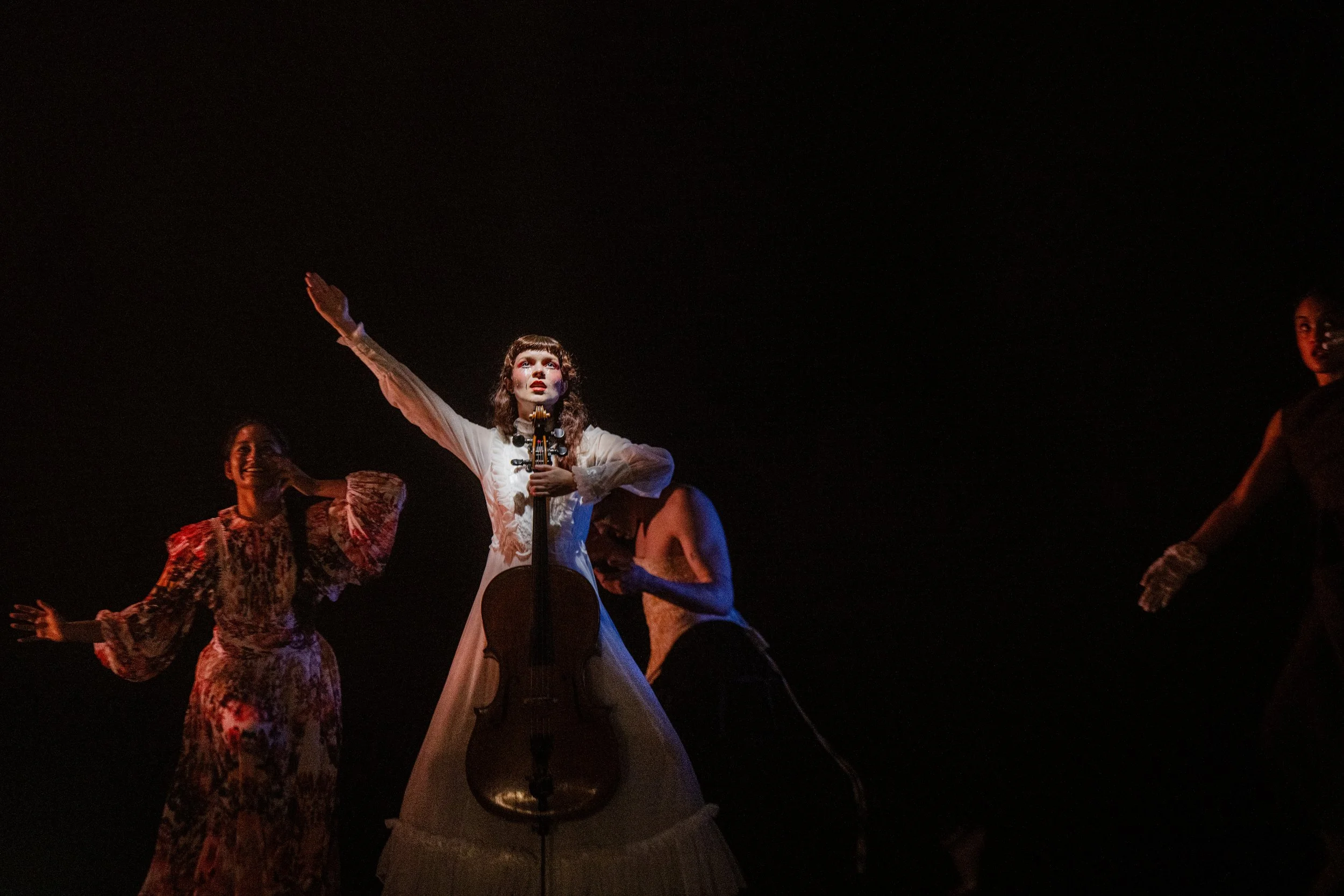 The image shows a theatrical performance featuring a woman in a white vintage dress with a cello at her waist, standing on stage with her arm raised, and three women in vintage-style dresses and gloves around her, all under dramatic lighting against 