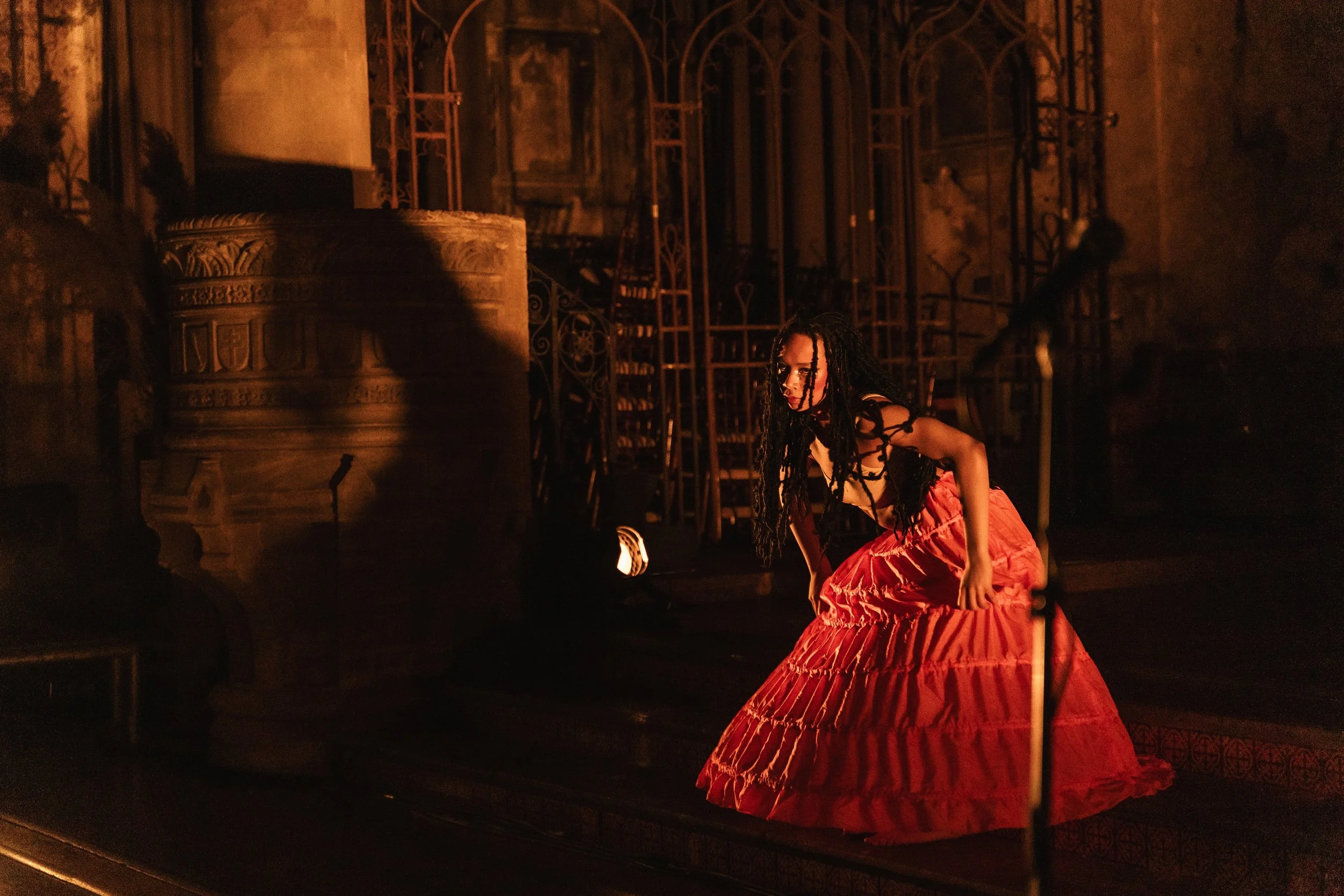A woman with dreadlocks wearing a black top and long red skirt on a dimly lit stage with ornate architectural features and wrought iron stairs.