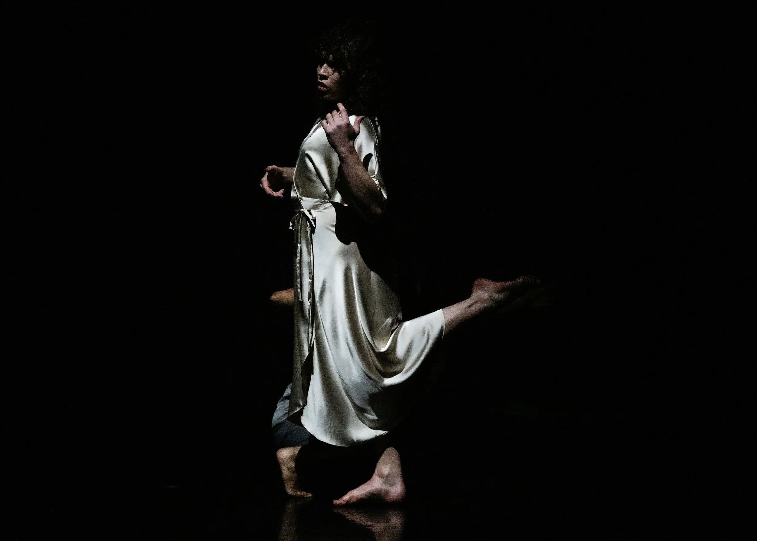 A woman in a silk dress kneels on one knee in a dark studio, with a black background, illuminated by a soft light highlighting her face, hair, and dress.
