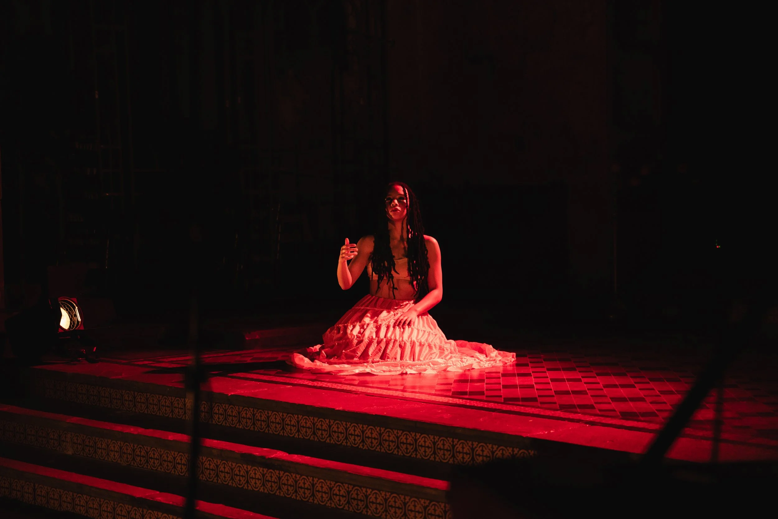 A woman with long, dark hair sitting on a stage illuminated by red light, dressed in a flowing, ruffled orange skirt and a matching top, with a dark background.