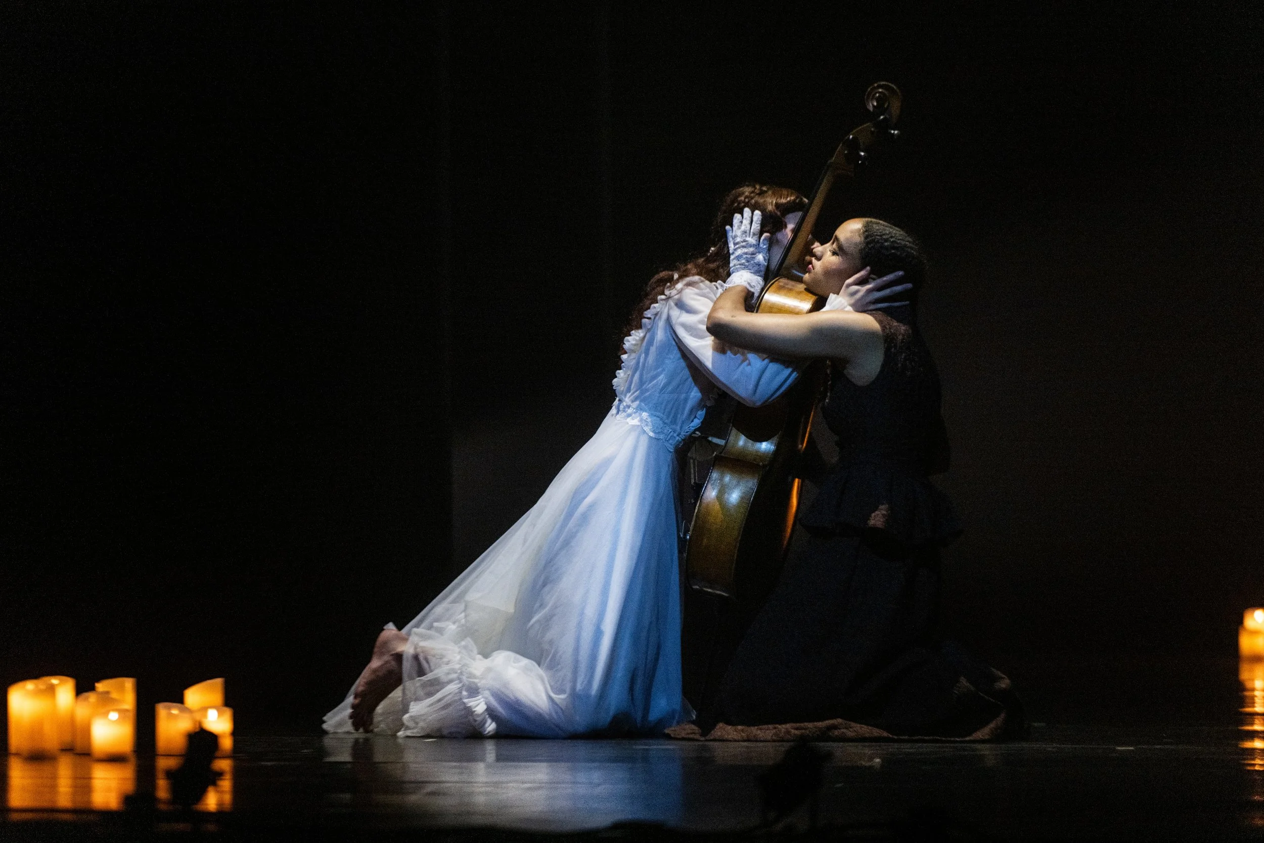 Two women, one in a white dress and the other in black, are kneeling and holding each other intimately in a dark setting with candles nearby.