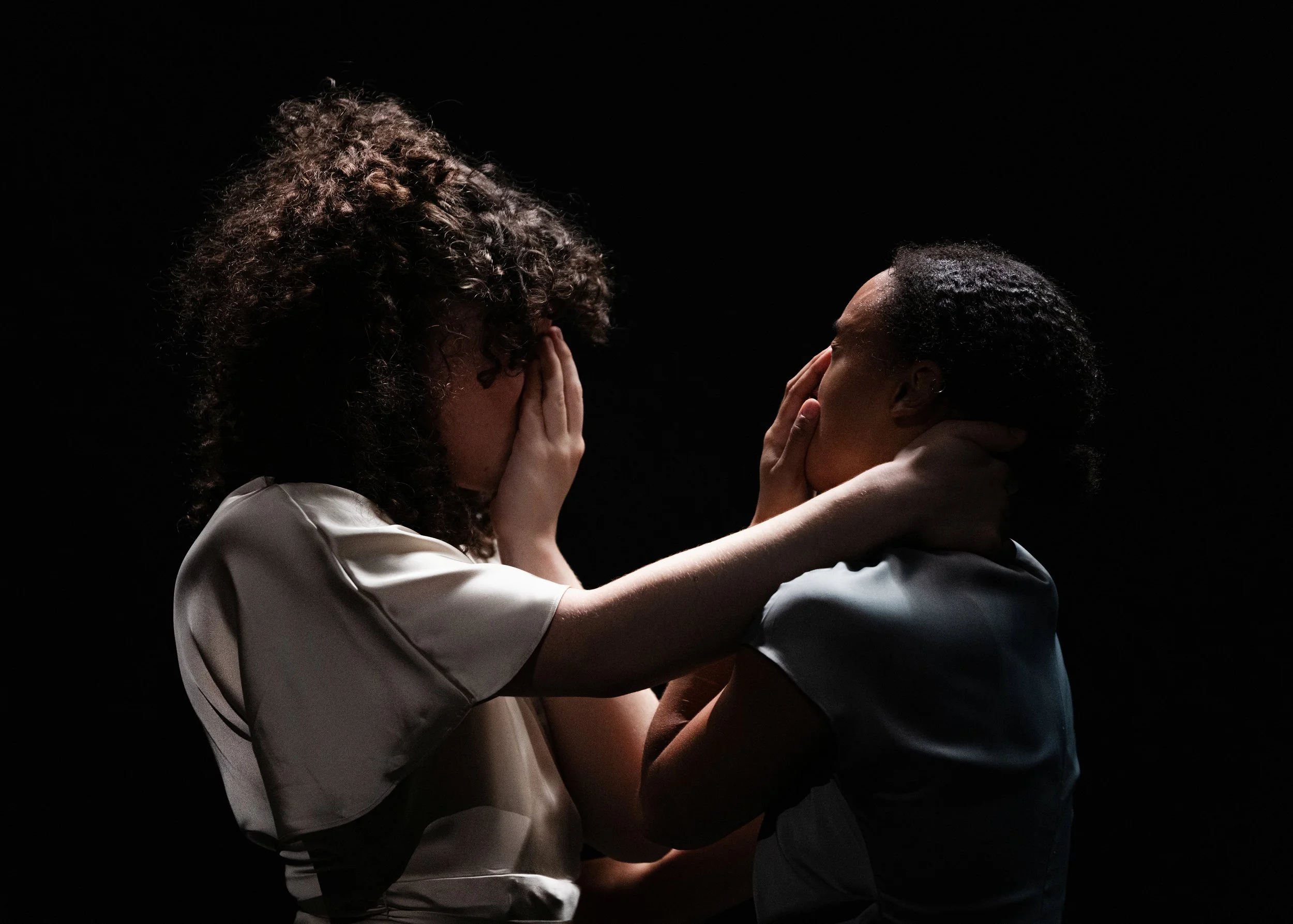 Two women face each other in an emotional moment, holding their faces and necks gently, with a black background.