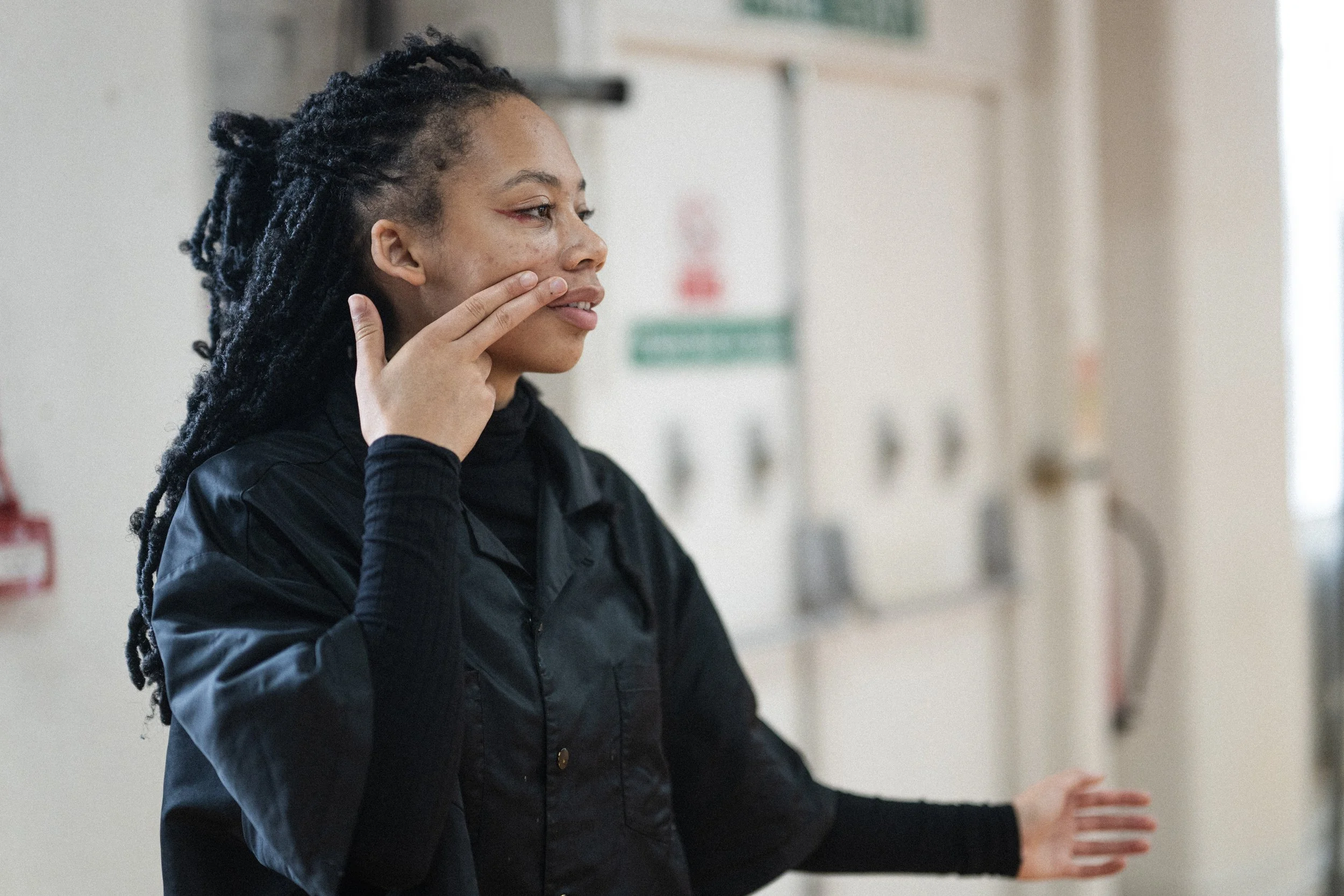 A woman wearing a black shirt is touching her face and smiling gently, standing in a room with a white wall and emergency signs in the background.