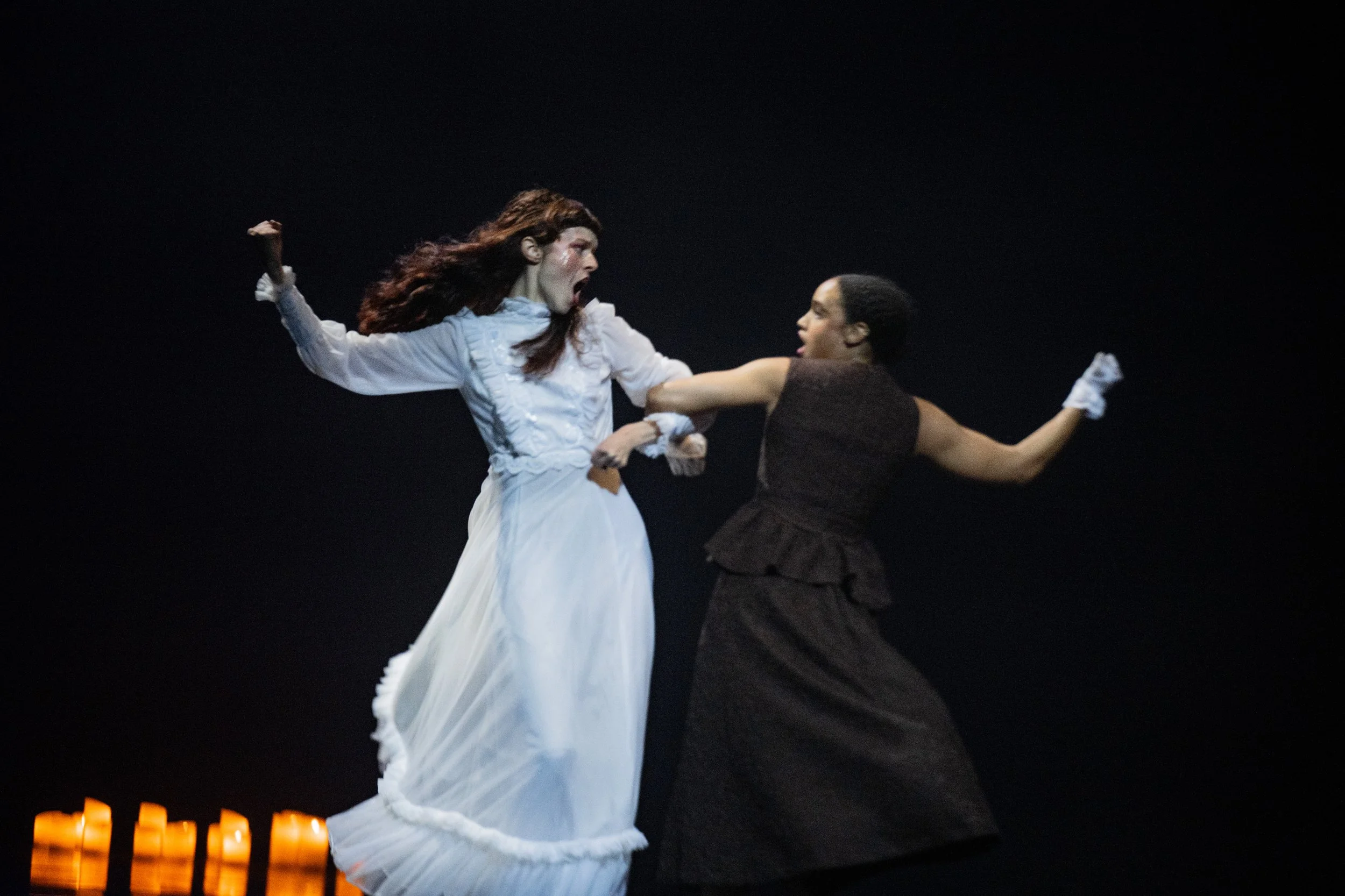 Two women in period costumes performing a theatrical scene on stage, with candles in the background.