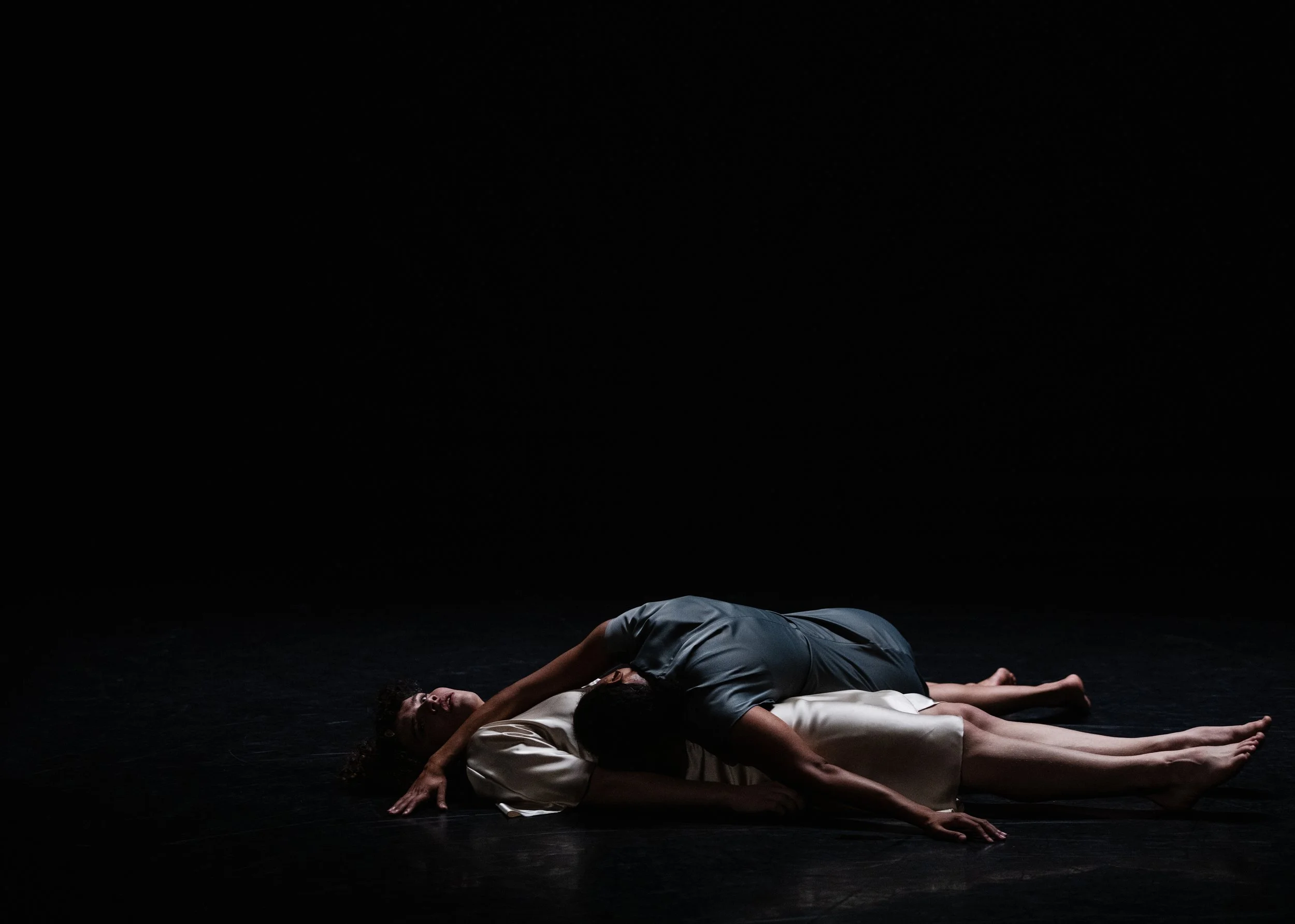 Two people lying on the floor in a dark room, one on top of the other, in an intimate pose.