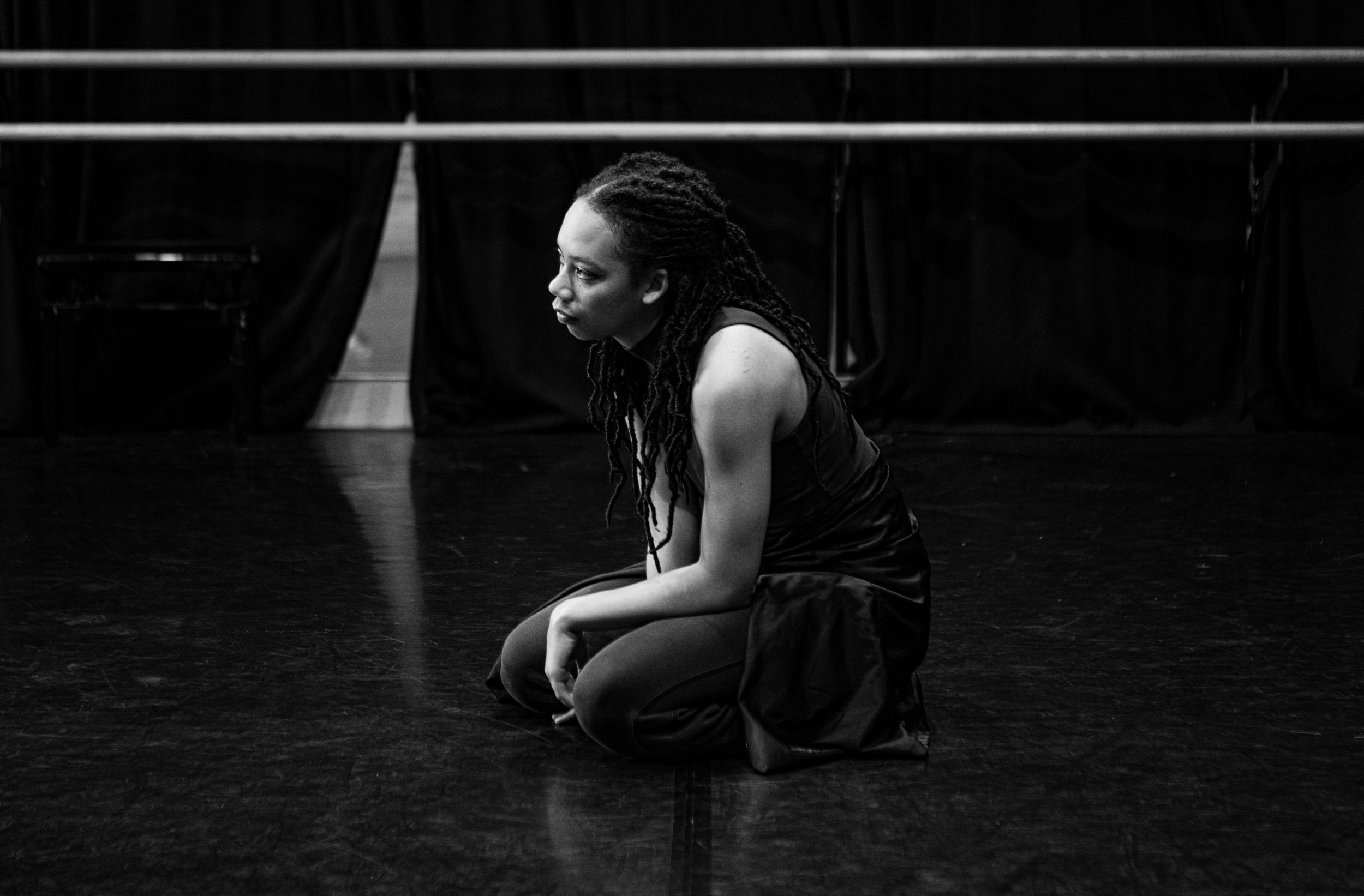 A woman kneeling in a dance studio appearing focused or contemplative.