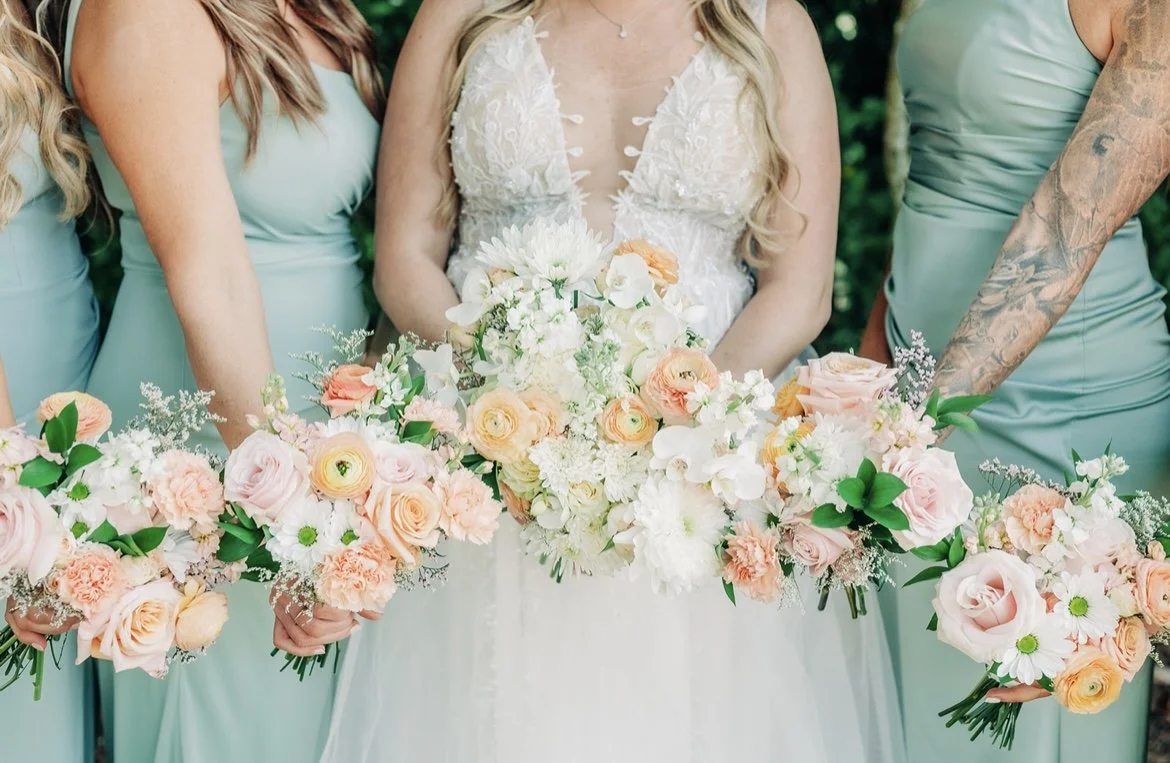 A bride and bridesmaids holding bouquets of pink, peach, and white flowers at a wedding.