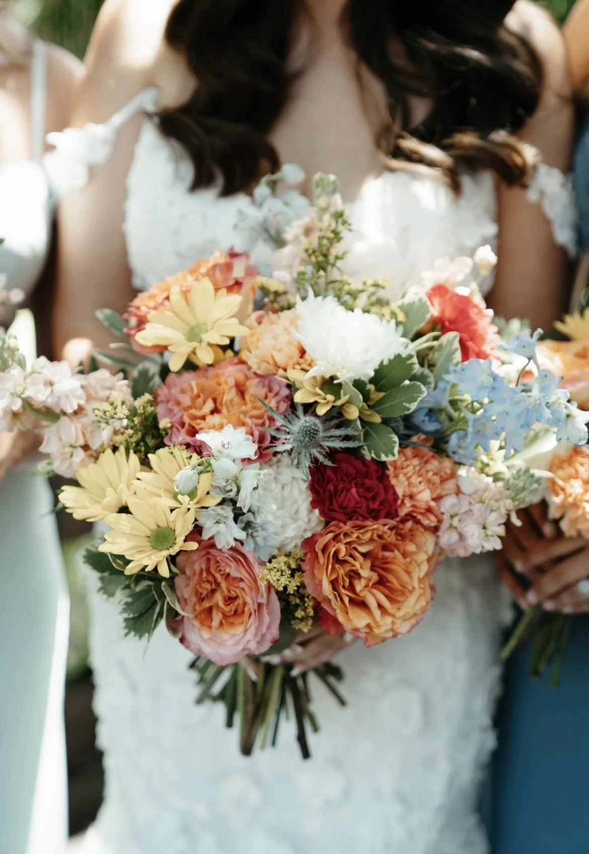 A person holding a large bouquet of colorful flowers, including roses, daisies, and other blossoms, in front of their white lace dress.