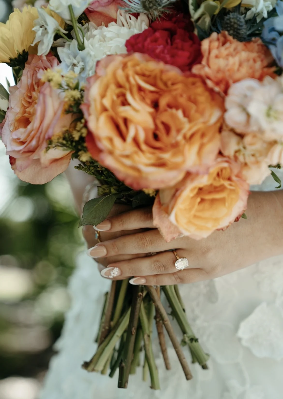 A person holding a bouquet of peach, pink, white, and red roses, with visible wedding rings on their fingers and manicured nails with white floral designs.