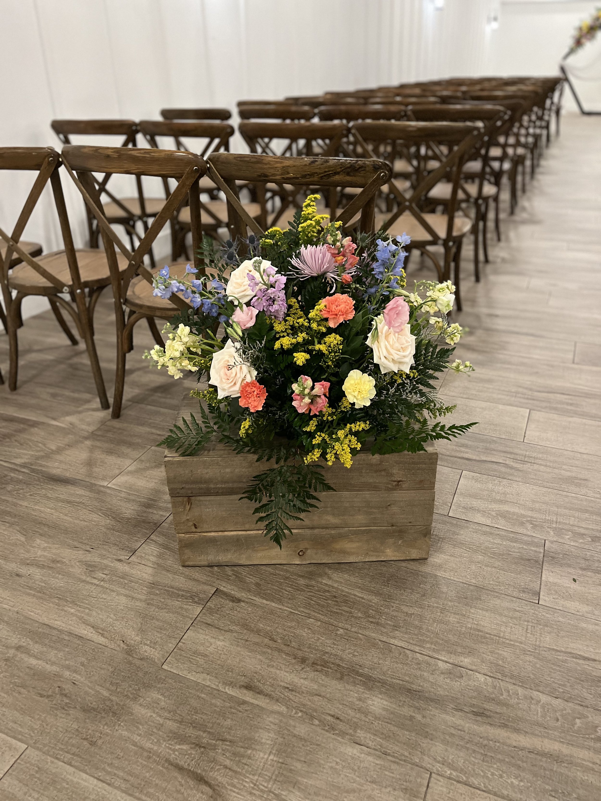 A floral arrangement in a wooden box with pink, white, yellow, purple, and peach flowers, some greenery, placed on a wooden floor with empty wooden chairs in the background.