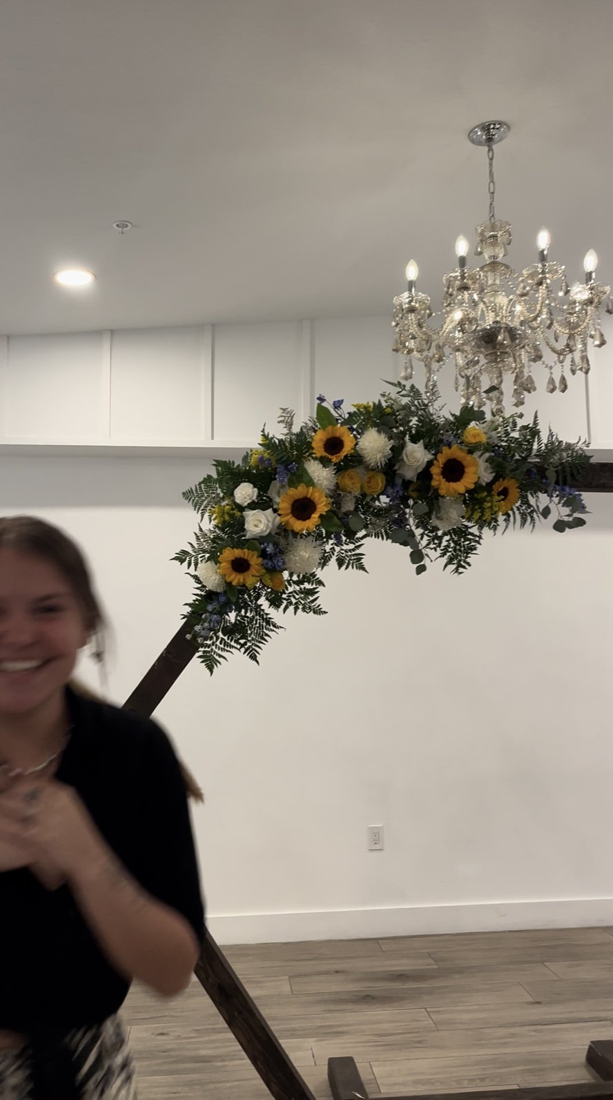 A woman smiling and partially visible on the left side of the image, standing next to a large floral arrangement decorated with sunflowers, white roses, and greenery, suspended from a wooden stand. A sparkling chandelier hangs from the ceiling above the floral display in a brightly lit room with a plain white wall and wooden flooring.