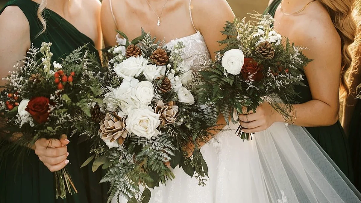 Close-up of three women holding large, festive wedding bouquets with white roses, red flowers, pinecones, and greenery, during a wedding celebration.
