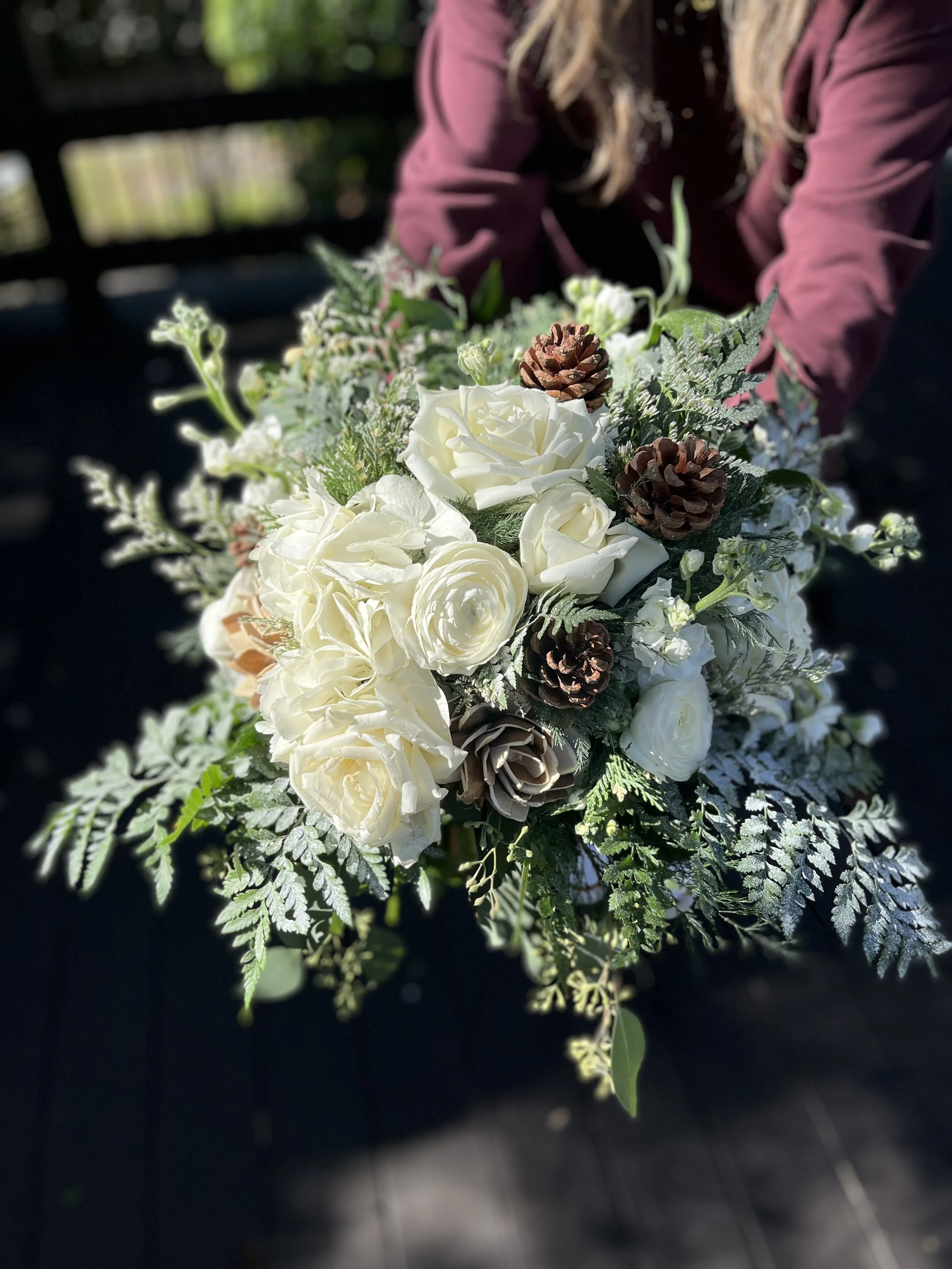 A person holding a bouquet of white roses, pinescones, and assorted greenery outdoors.