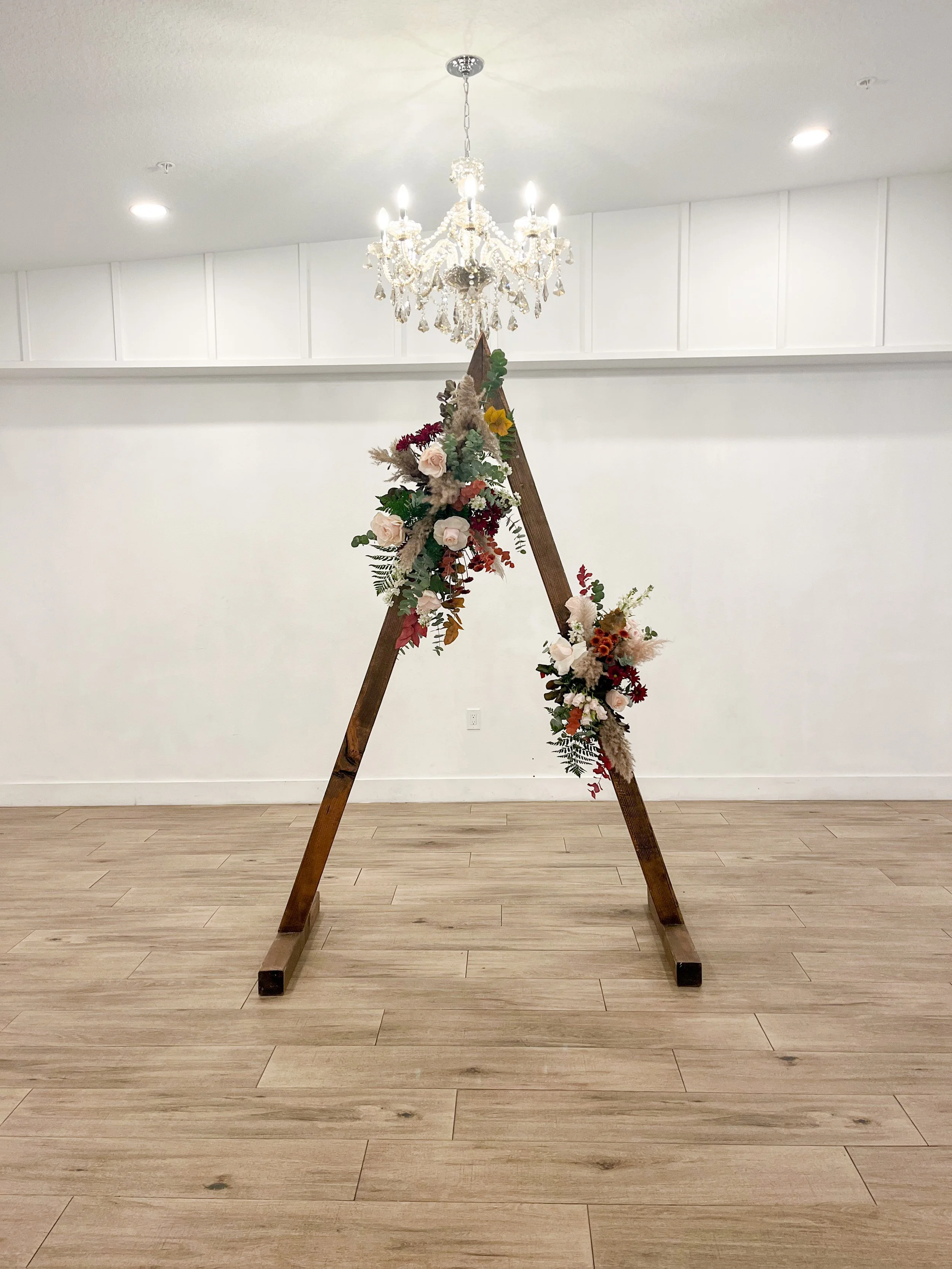 A wooden A-frame stand decorated with flowers, placed on a light wood floor in a room with white walls and a chandelier overhead.