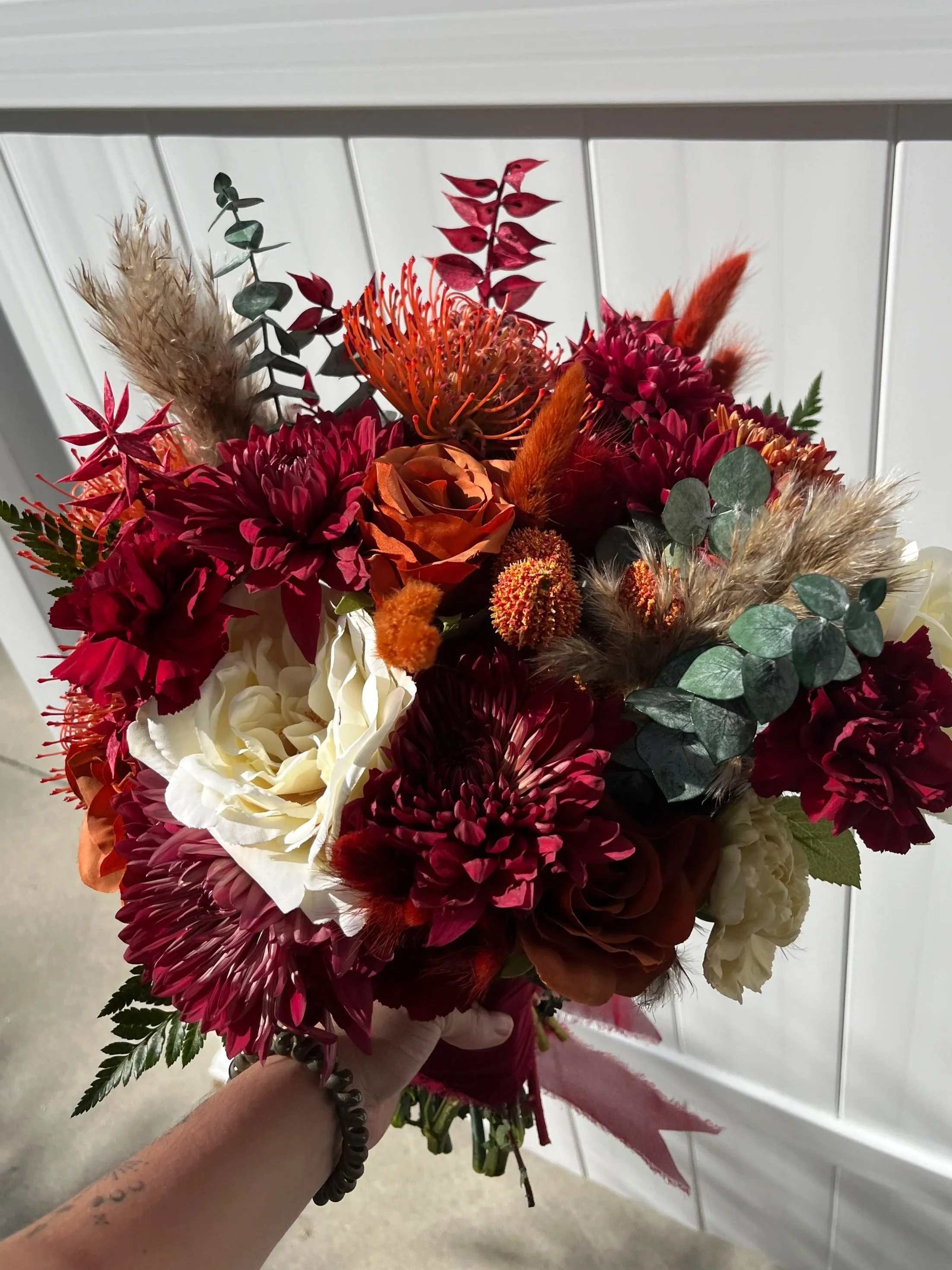 A colorful bouquet of mixed flowers including burgundy, white, orange, and pink blooms with greenery and soft feathers, held by a person with a visible tattoo on their wrist.