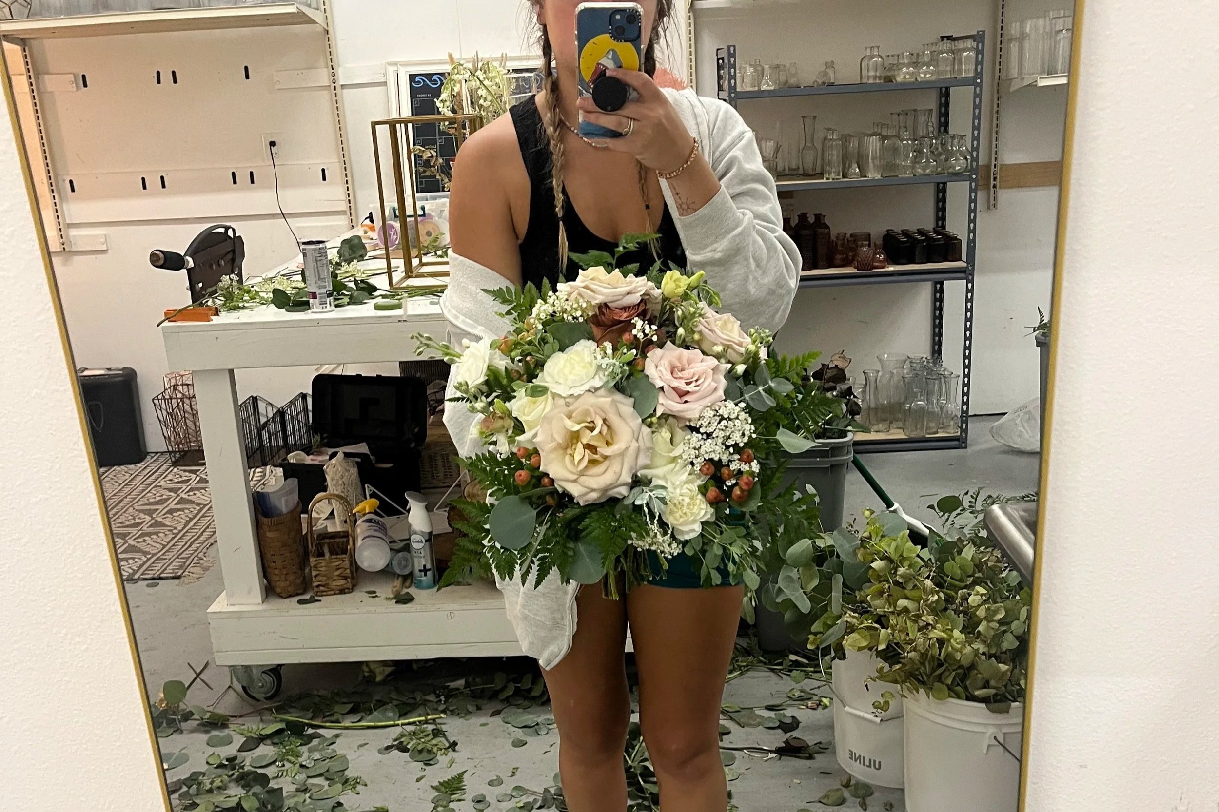 Person taking a mirror selfie holding a large bouquet of flowers in a room with shelves and assorted items.